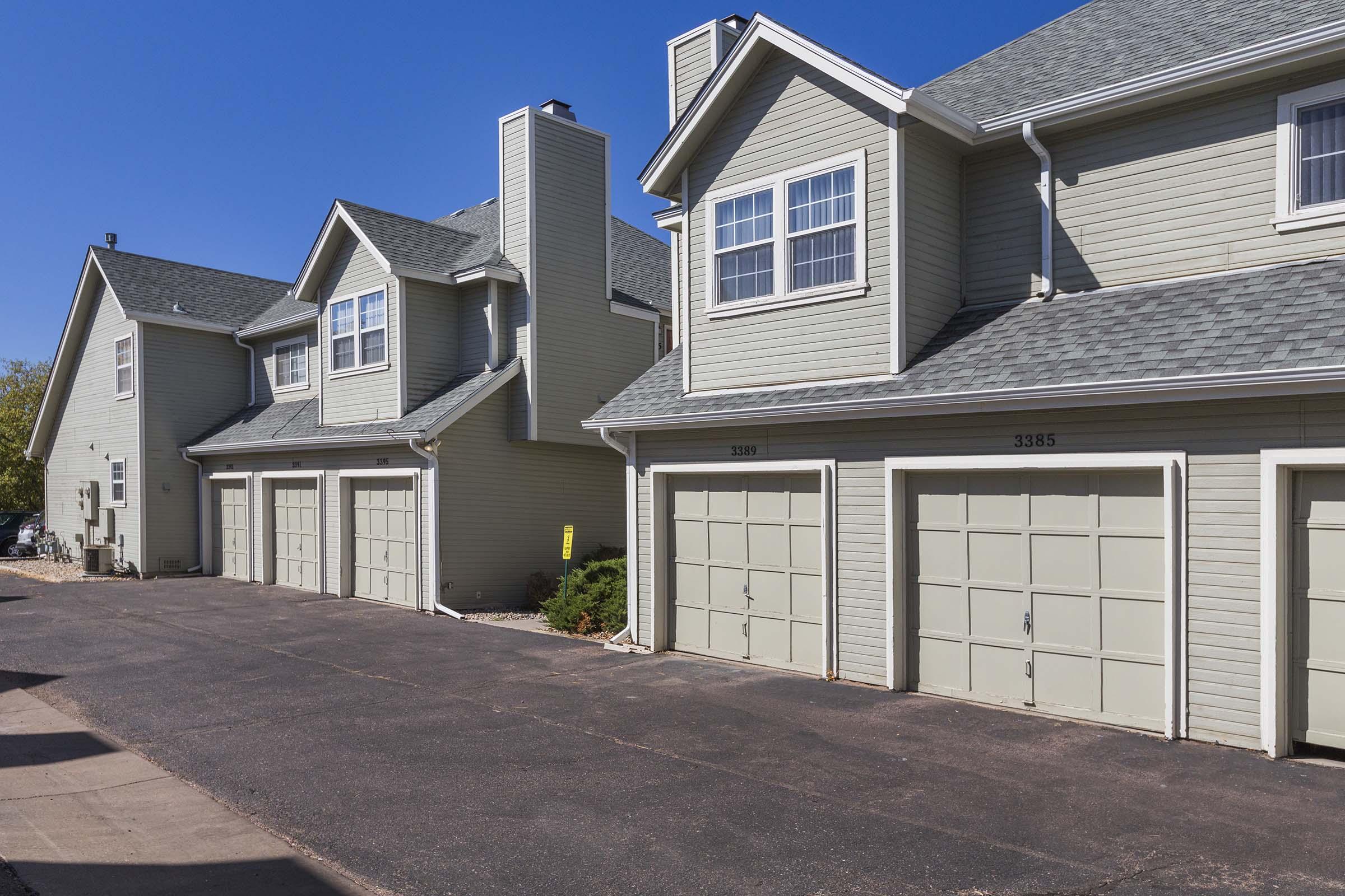 A row of residential buildings featuring several garages with beige siding, a clear blue sky overhead, and a paved driveway. The garages have light-colored doors, and the area is well-maintained with some shrubs visible near the buildings.