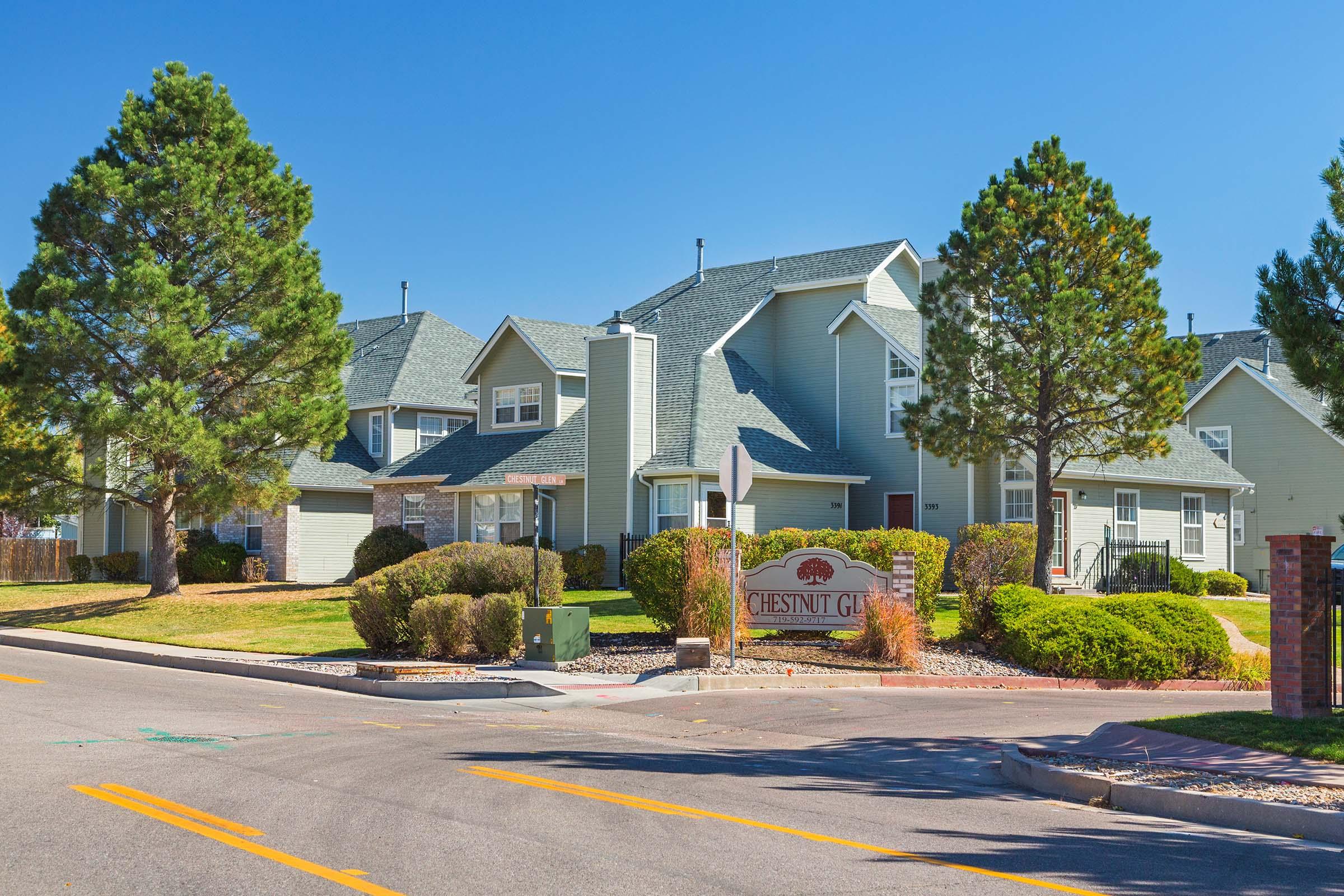 A view of a residential neighborhood featuring modern houses with gabled roofs, surrounded by trees and landscaped lawns. A sign reading "Chestnut Glen" is prominently displayed at the entrance, and the scene is set under a clear blue sky.