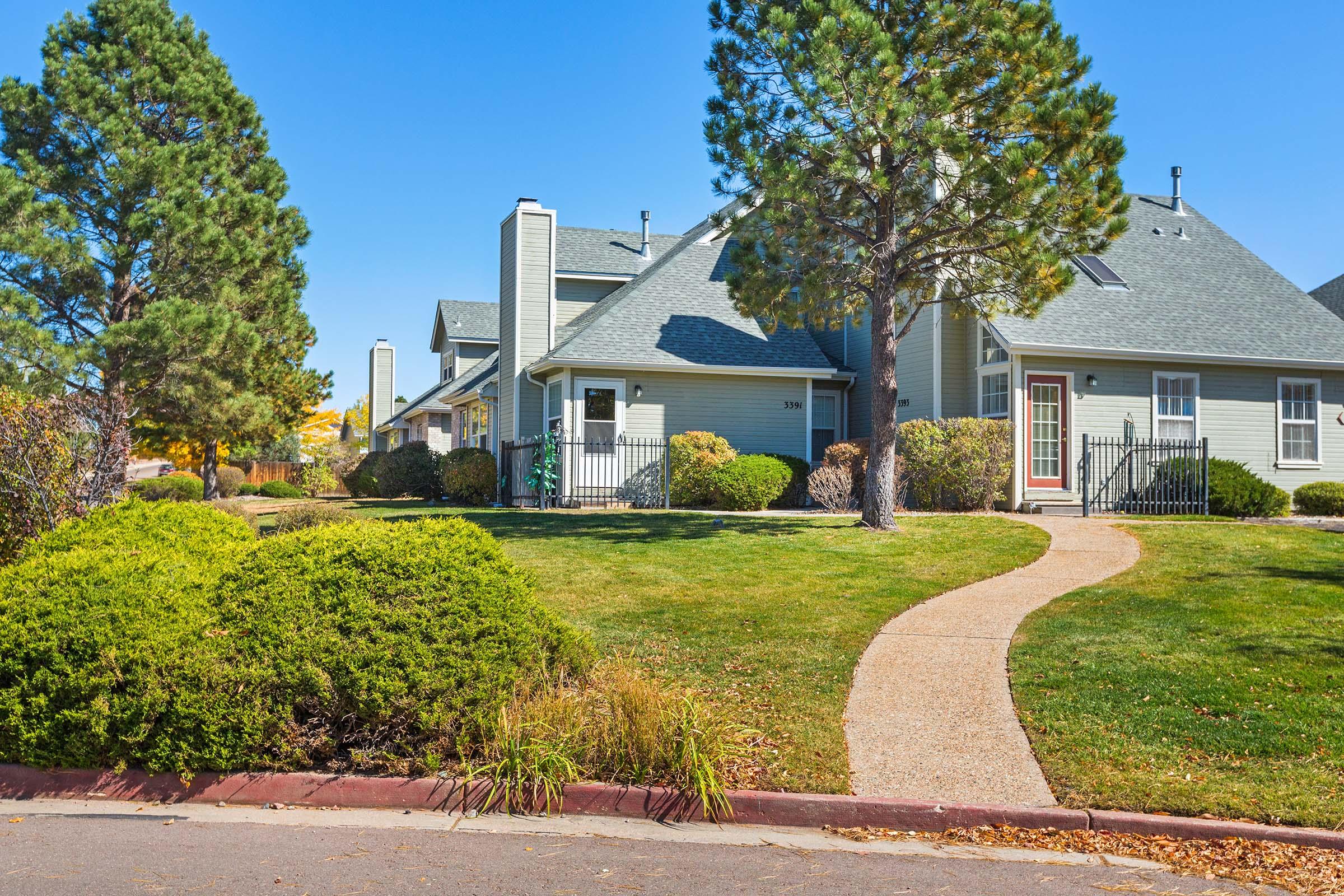 A well-maintained residential area featuring two-story houses with gray roofs. A winding pathway leads through a green lawn with neatly trimmed bushes and trees. Bright blue sky overhead, indicating clear weather.
