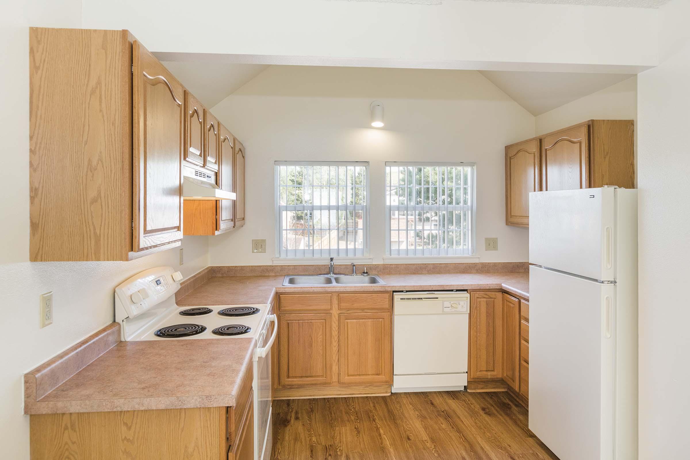 Bright kitchen featuring wood cabinetry, a countertop with a sink, and appliances including an oven, dishwasher, and refrigerator. Large windows allow natural light to illuminate the space, which has a light-colored wall and a wooden floor.