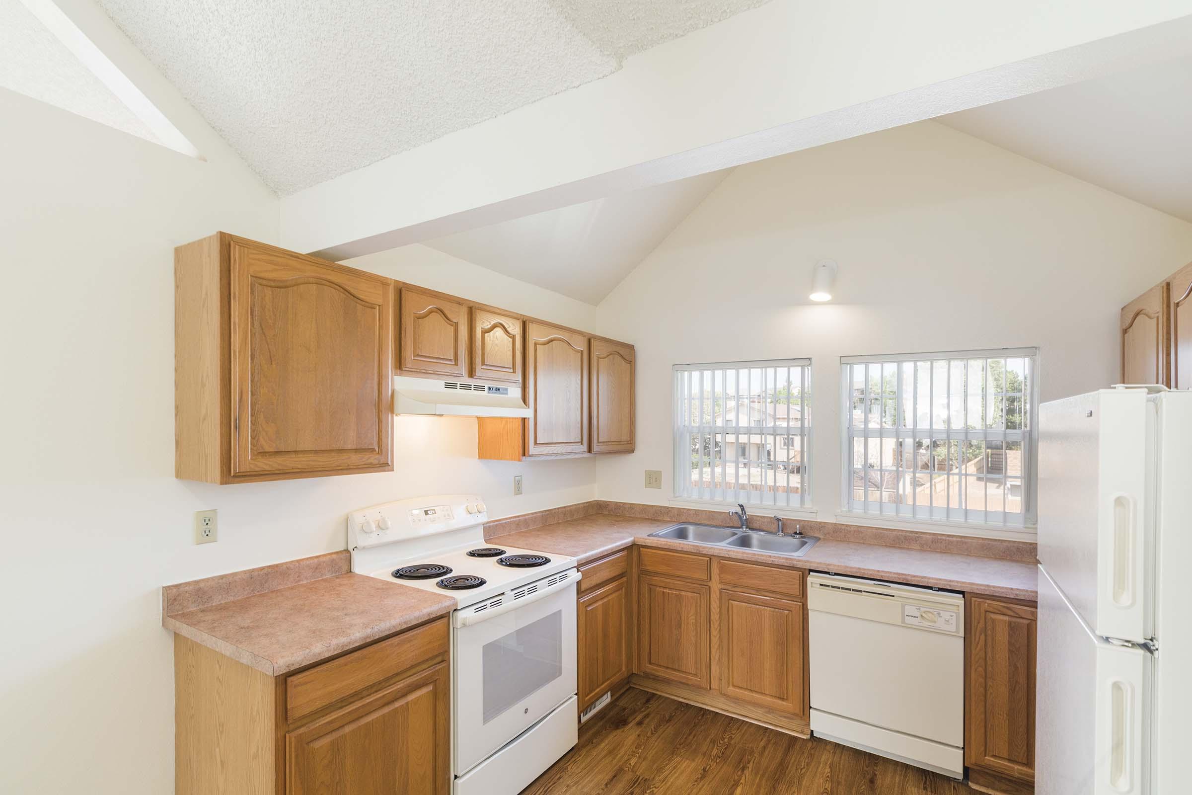 Bright kitchen with wooden cabinetry, a white stove, refrigerator, and dishwasher. Features a sink beneath two windows, allowing natural light to illuminate the space. The ceiling is high with a light fixture, and the flooring is a warm wood finish.