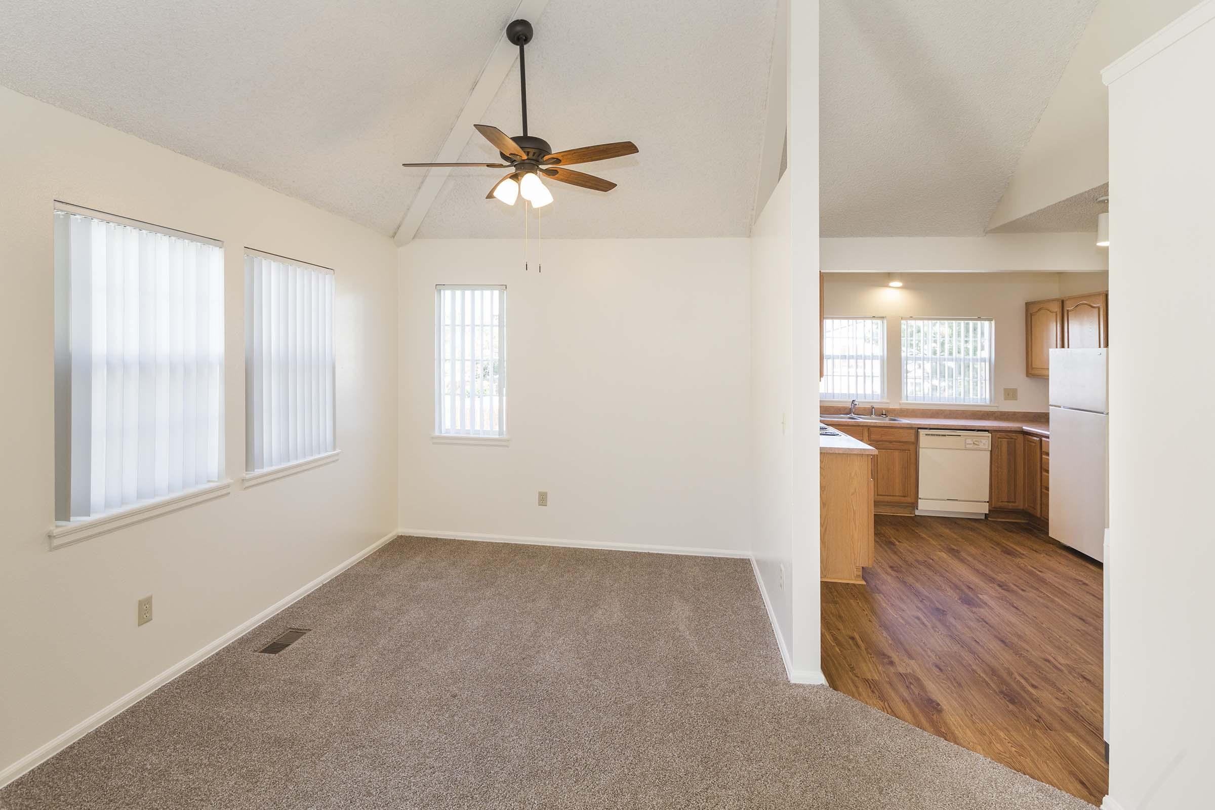 Spacious living area featuring a ceiling fan, light-colored walls, and large windows allowing natural light. The open layout connects to a kitchen with wooden cabinets and appliances, complemented by warm wood flooring. Ideal for comfortable living and entertaining.