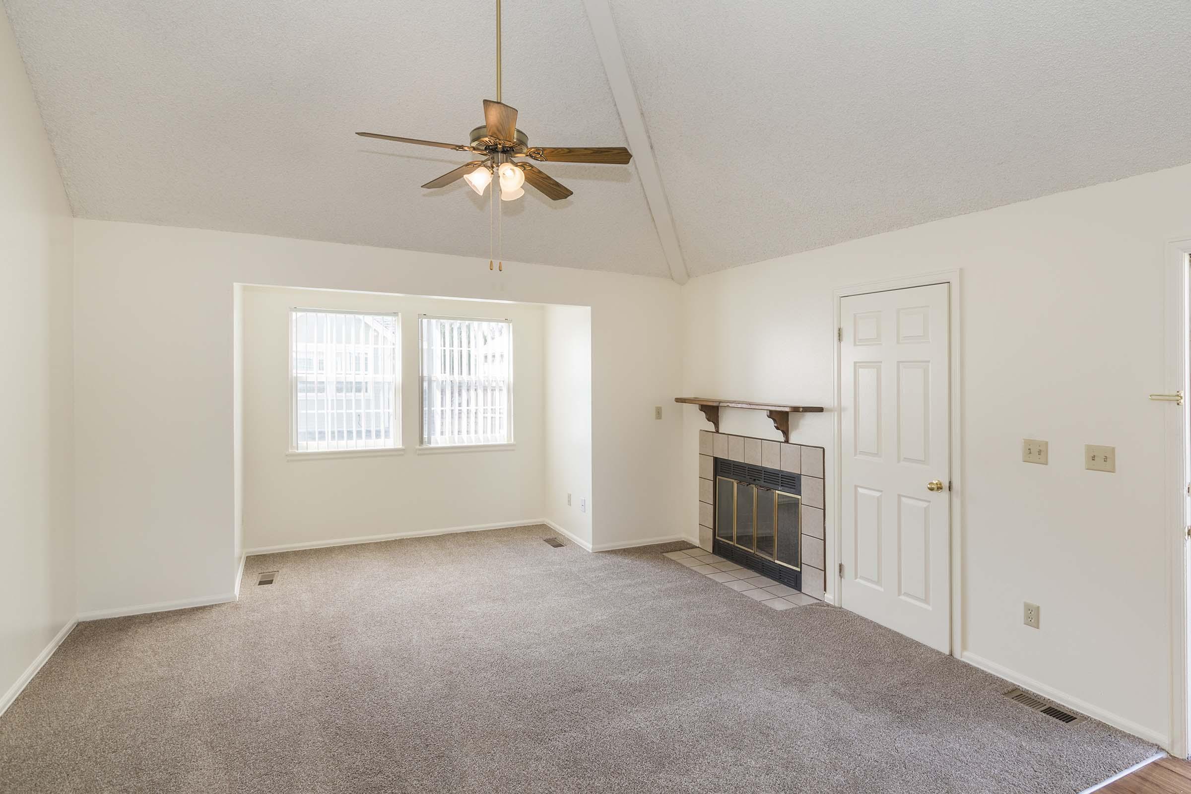 A spacious living room with light-colored walls and a ceiling fan. The room features large windows allowing natural light, a fireplace against one wall, and a door leading to another area. The floor is carpeted, and the ceiling has a peaked design, enhancing the room's openness.