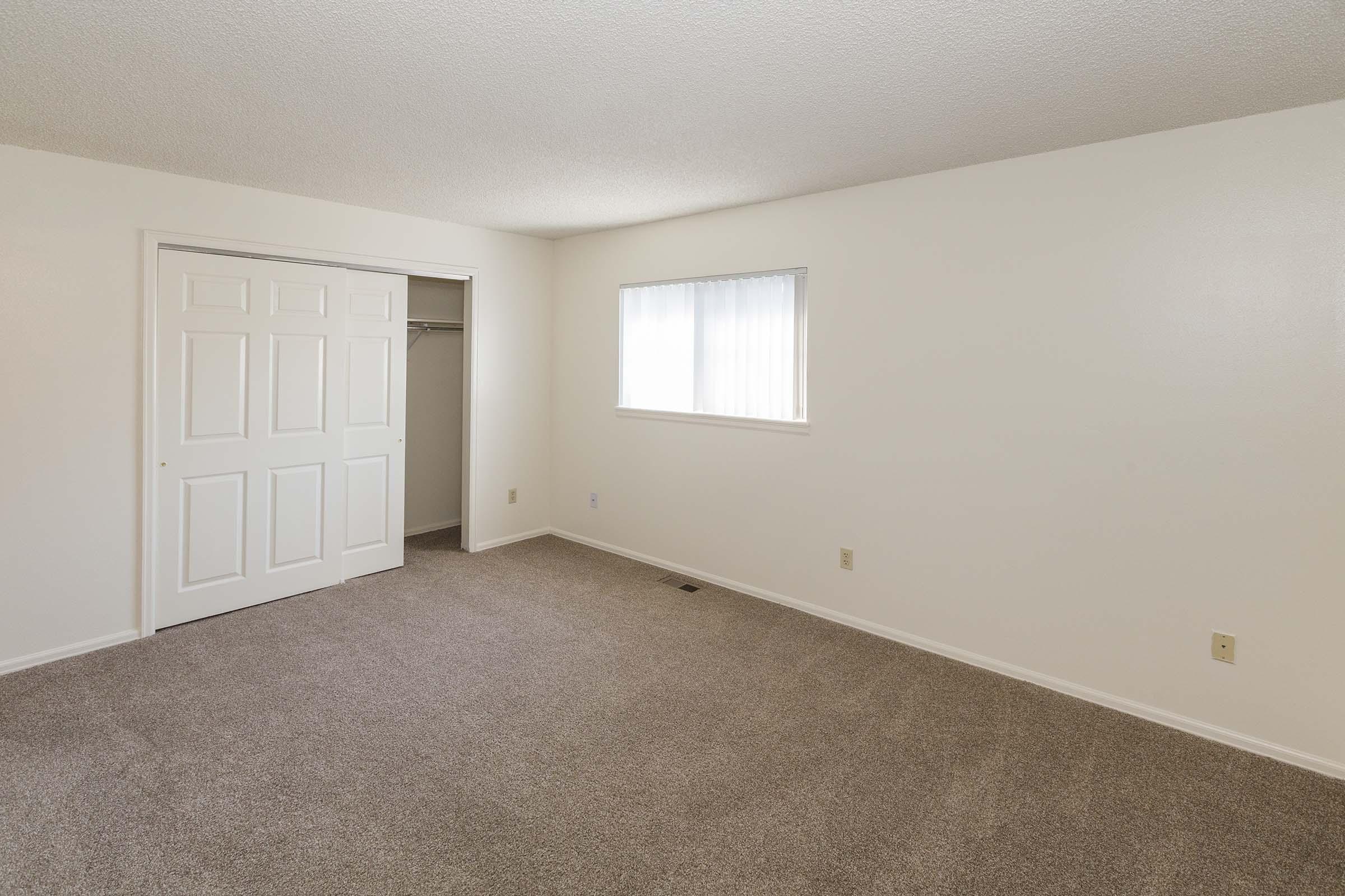 A spacious, empty room with light beige carpet, white walls, and a window letting in natural light. There is a closet with sliding doors on the left side, and the overall atmosphere is bright and airy, ideal for customizable interior decorating.