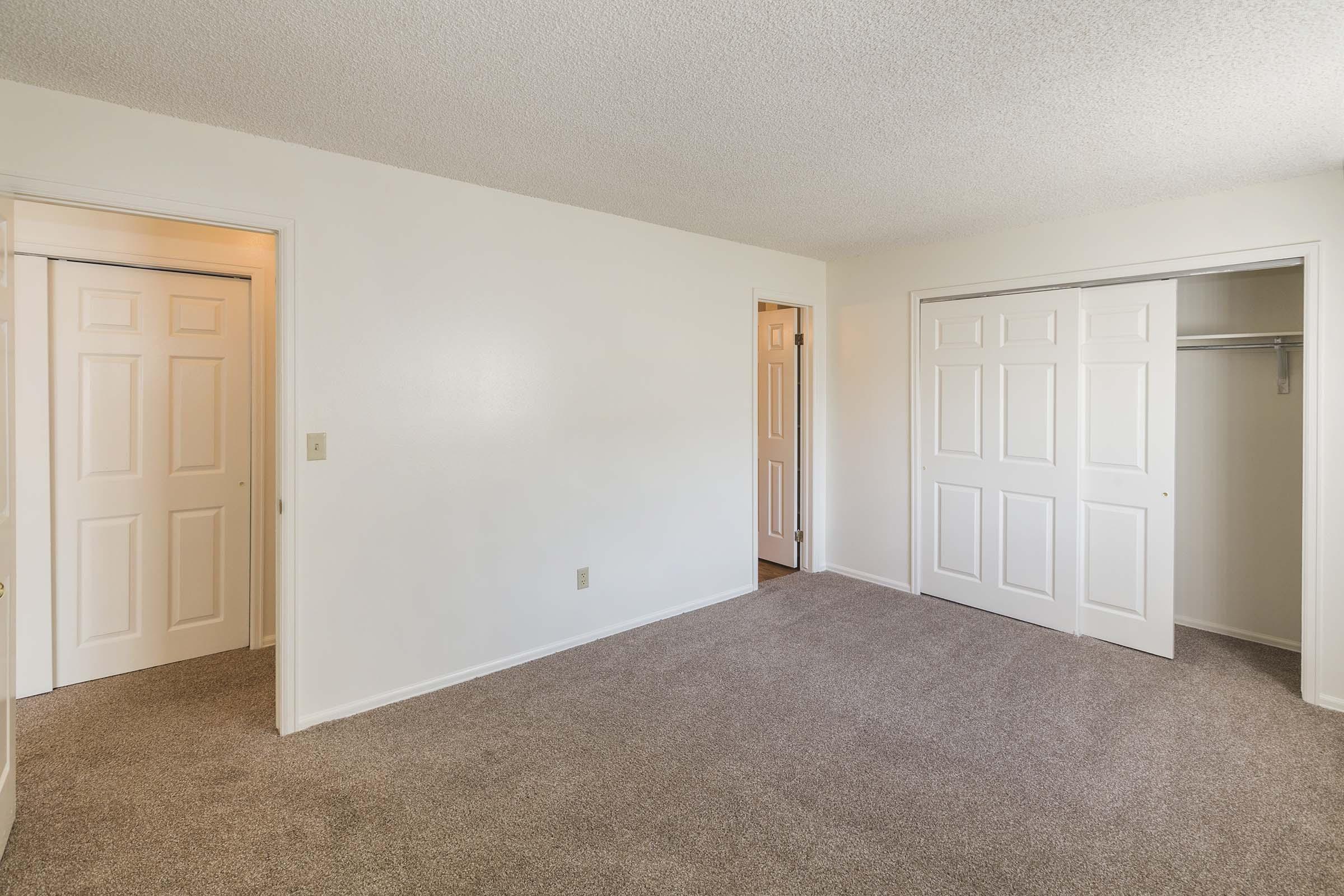 A spacious, empty room with light-colored walls and carpeted flooring. There are two doors on the left leading to closets, and another door on the right, providing access to another area. The room has a bright and neutral aesthetic, suitable for various uses.