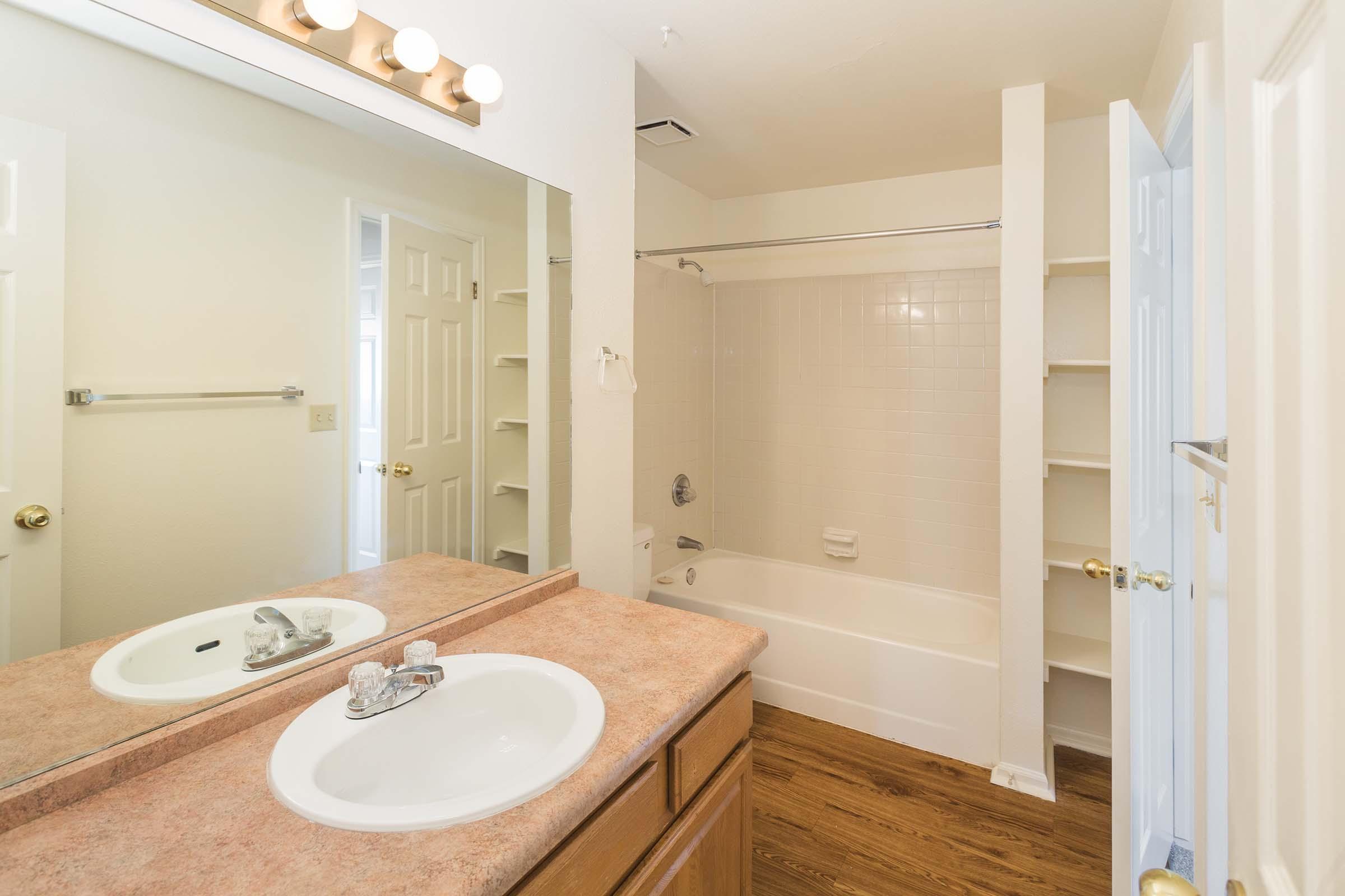 A bright bathroom featuring a vanity with a sink and mirror, a shower/tub combo, and light-colored walls. There are wooden cabinets below the sink and shelves for storage. The flooring is a warm wood tone, enhancing the overall spacious and clean appearance.