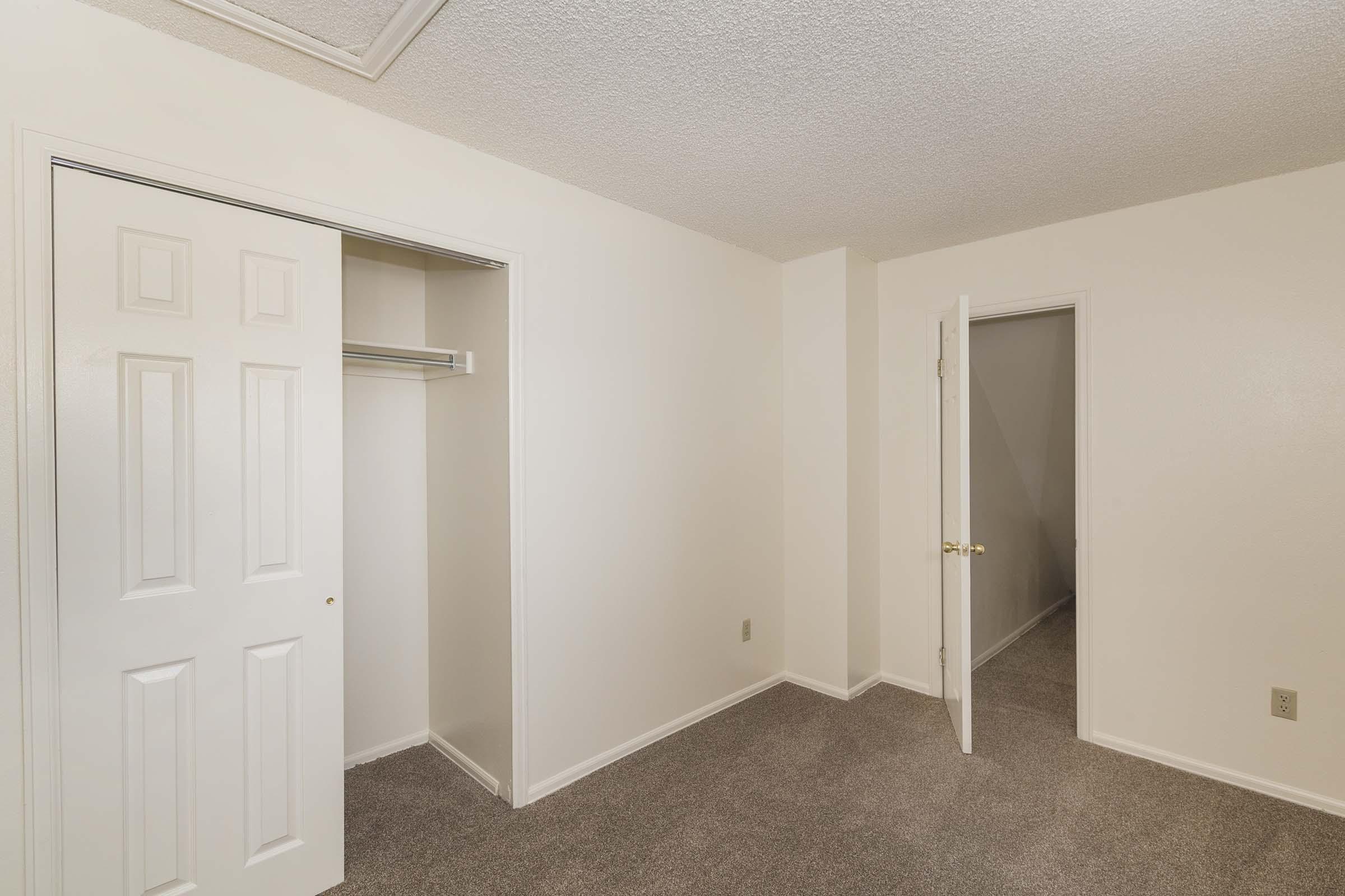 A small, empty room featuring light-colored walls and beige carpeting. There are two doors: one leading to a closet with a sliding door and another door opening to a hallway. The ceiling has a textured finish, and the overall ambiance is bright and minimalistic.