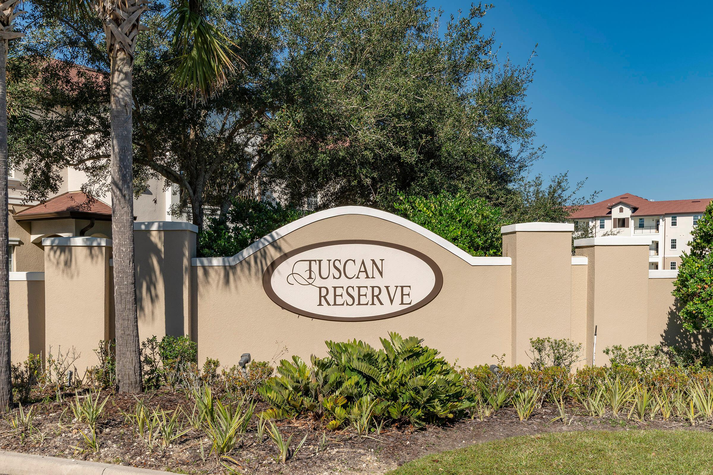Sign for Tuscan Reserve, set against a clear blue sky. The sign is situated in front of a landscaped area with shrubs and palm trees, and is part of a residential community featuring low-rise buildings in the background.