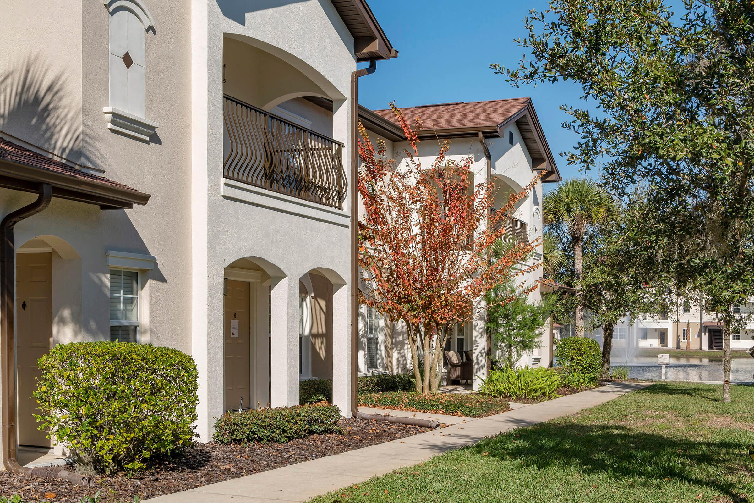 Two multi-story residential buildings with white stucco exteriors and brown roofs. One building features a balcony, and both are surrounded by well-maintained landscaping, including bushes and a small tree with red leaves. The scene is bathed in sunlight, indicating a clear, sunny day.