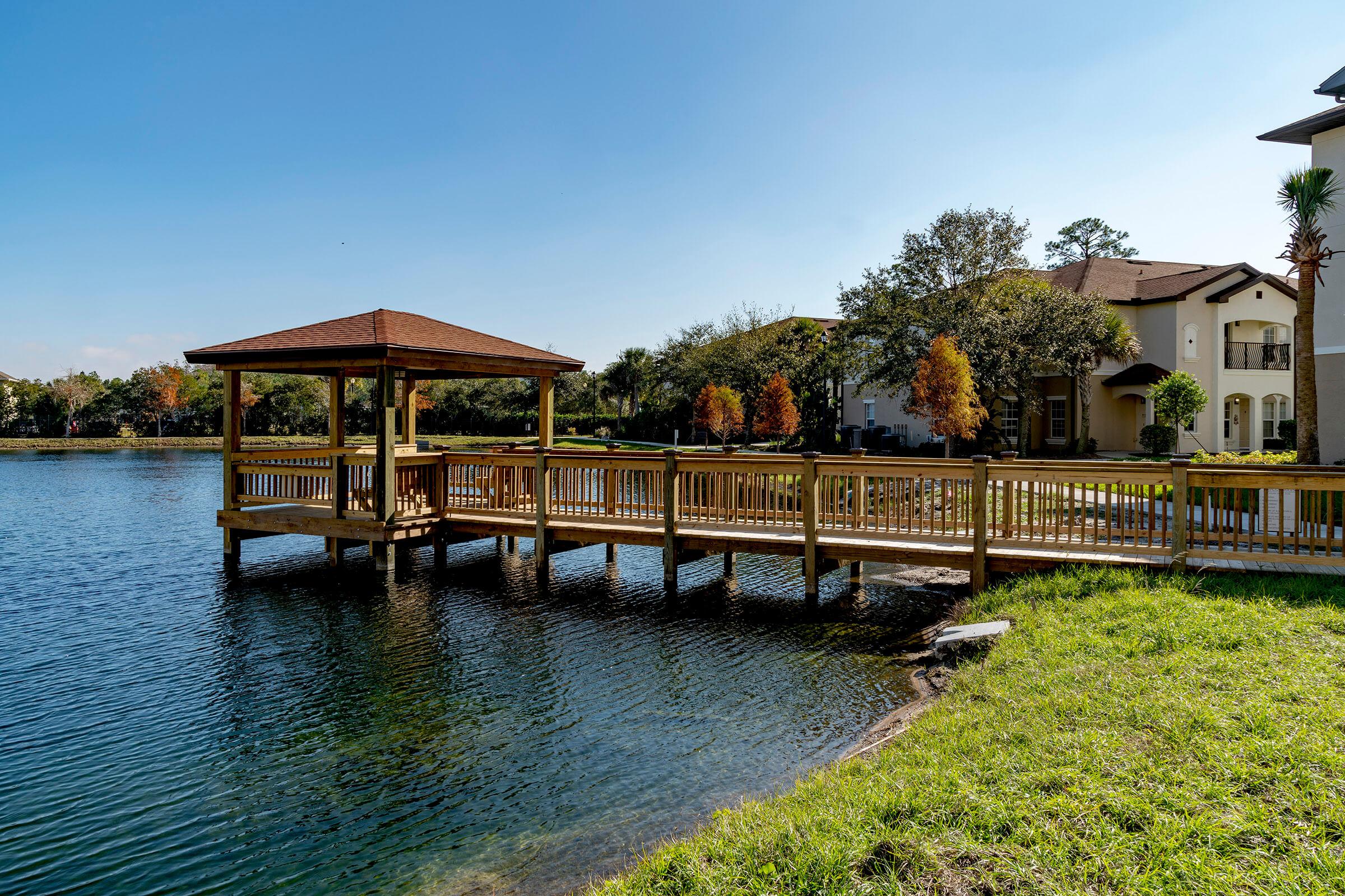 A wooden gazebo on a dock extends over a calm lake, surrounded by vibrant trees with autumn colors. In the background, residential buildings can be seen against a clear blue sky. The scene captures a peaceful waterfront setting, perfect for relaxation and contemplation.