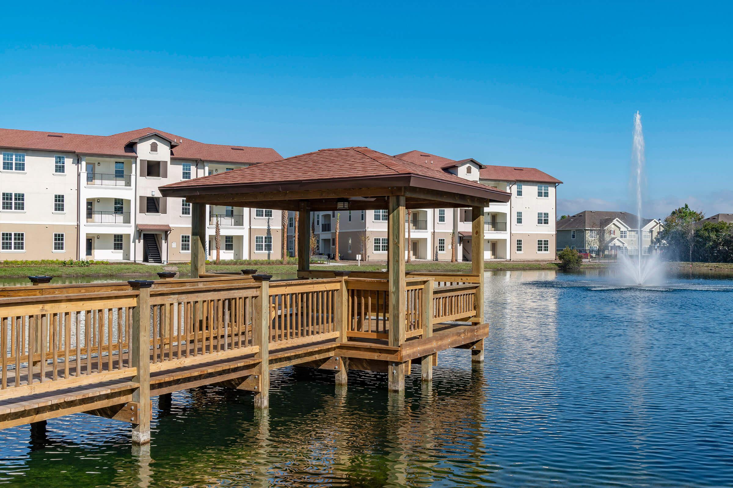 A wooden gazebo extends over a calm pond, surrounded by greenery. In the background, several residential buildings with red roofs are visible. A fountain in the pond adds a tranquil touch, creating a serene lakeside atmosphere. Clear blue skies complement the picturesque scene.