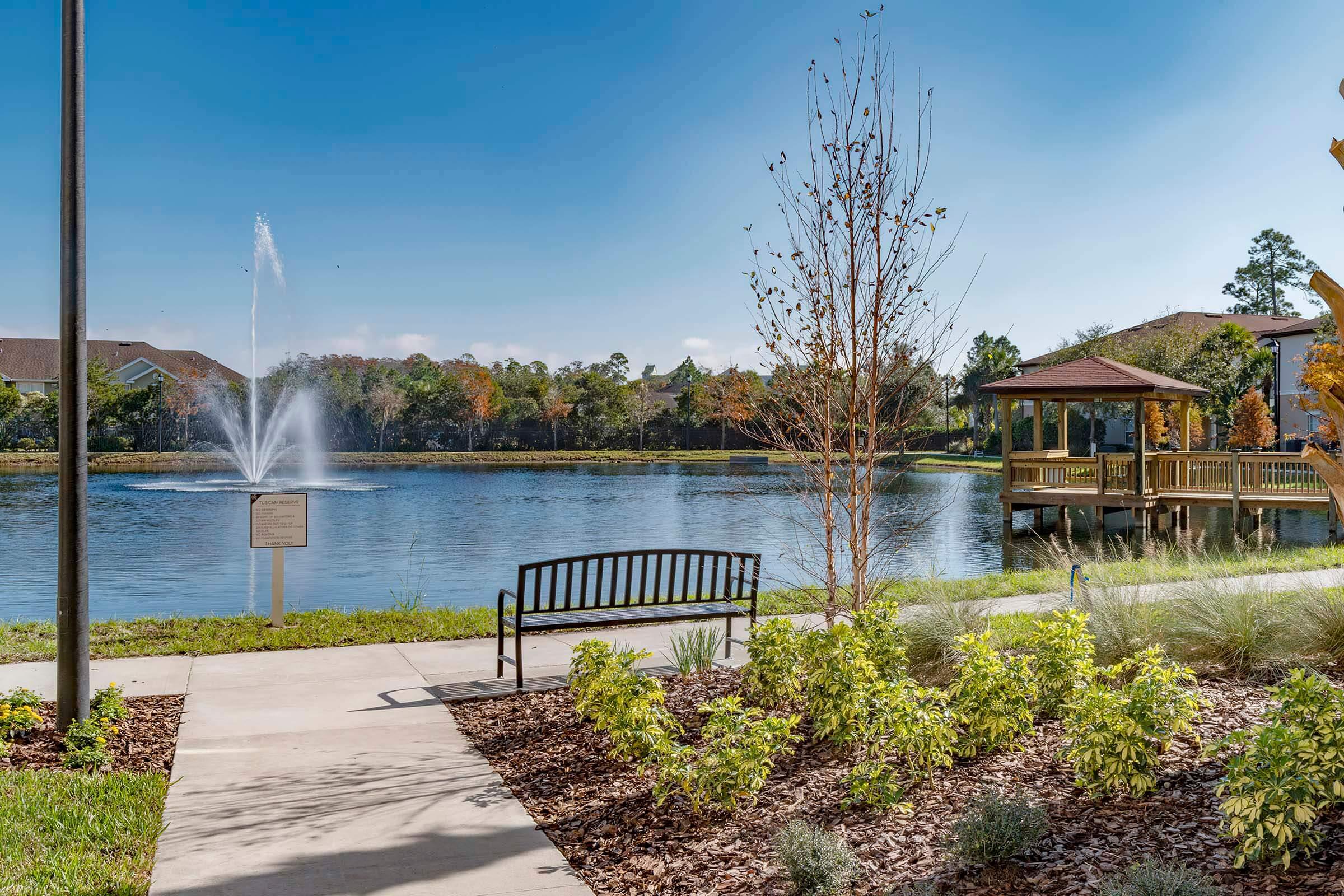 A serene park scene featuring a paved walkway leading to a bench overlooking a calm pond. A decorative fountain rises from the water, surrounded by lush greenery and colorful trees in the background. A gazebo can be seen nearby, adding to the tranquil atmosphere.