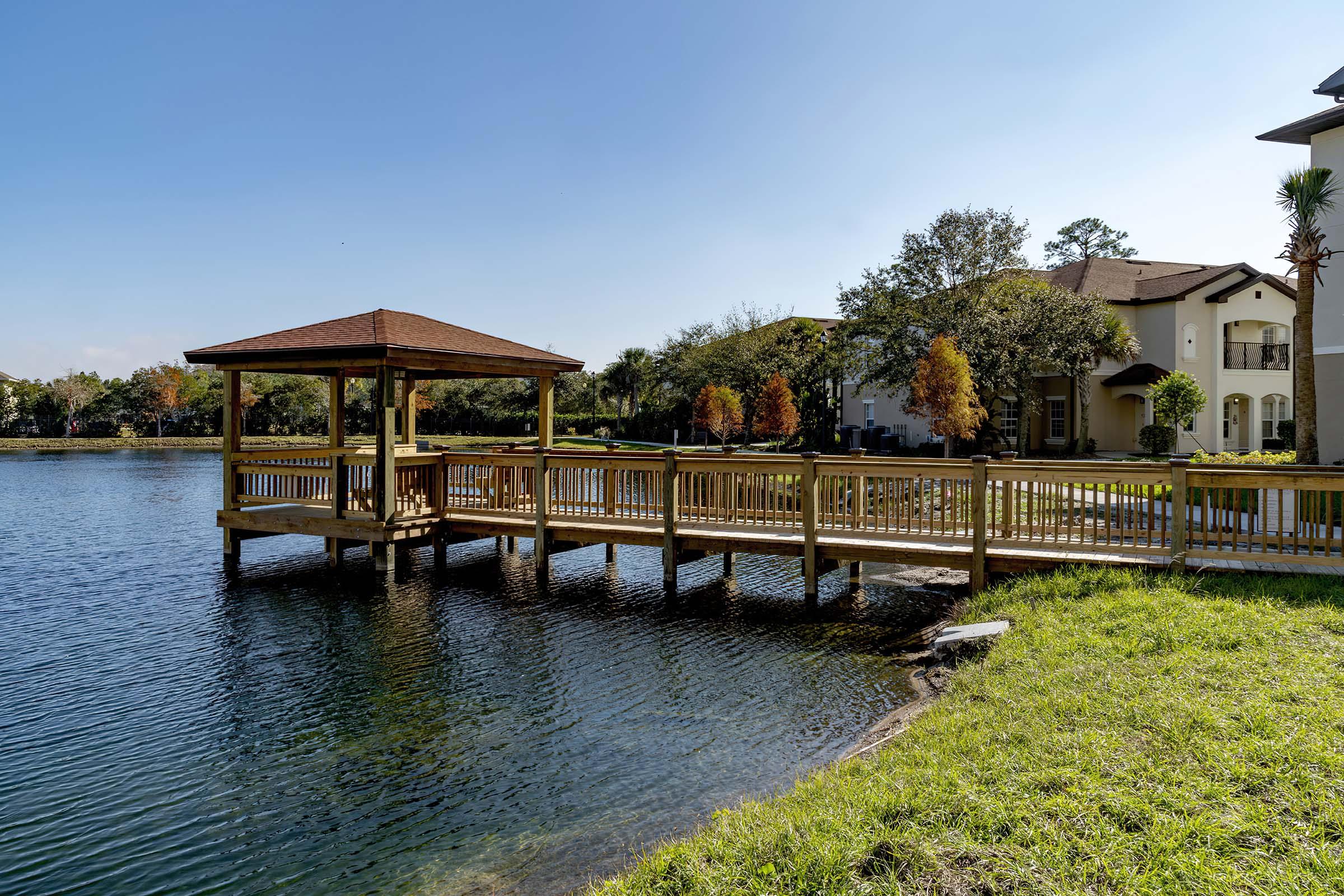 A wooden gazebo on a dock extends over a serene lake, surrounded by lush green grass and trees with autumn foliage. In the background, a residential building complements the tranquil setting under a clear blue sky.