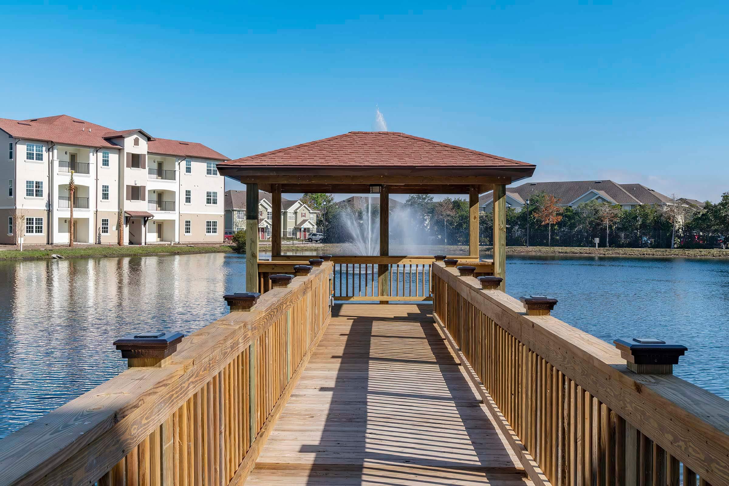 A wooden dock leading to a gazebo on a calm lake, surrounded by multi-story residential buildings. A fountain sprays water in the distance, under a clear blue sky. The scene conveys a peaceful outdoor setting, ideal for relaxation and leisure.