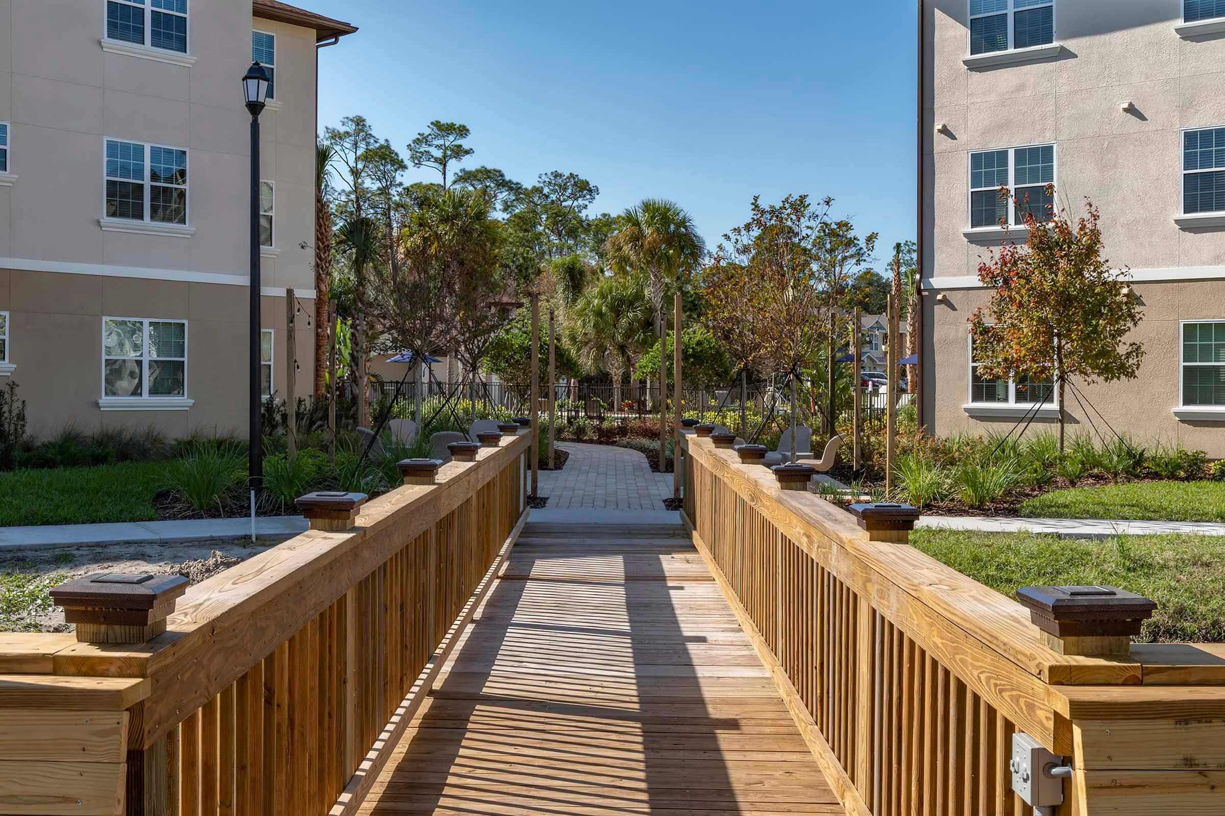 A wooden bridge leads through a landscaped area between two buildings. Lush greenery and trees are visible on either side, with paved pathways and seating areas. The sun shines brightly, creating a warm and inviting atmosphere in this outdoor space.