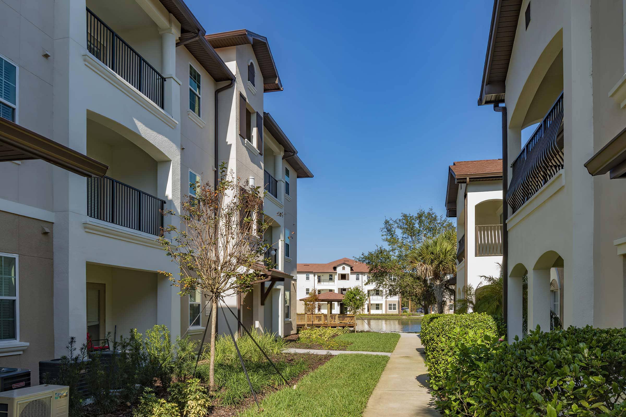 A well-maintained apartment complex with two buildings flanking a paved walkway. Green grass and small trees line the path, and clear blue sky is visible overhead. The complex features balconies and well-kept landscaping, creating a peaceful residential atmosphere.