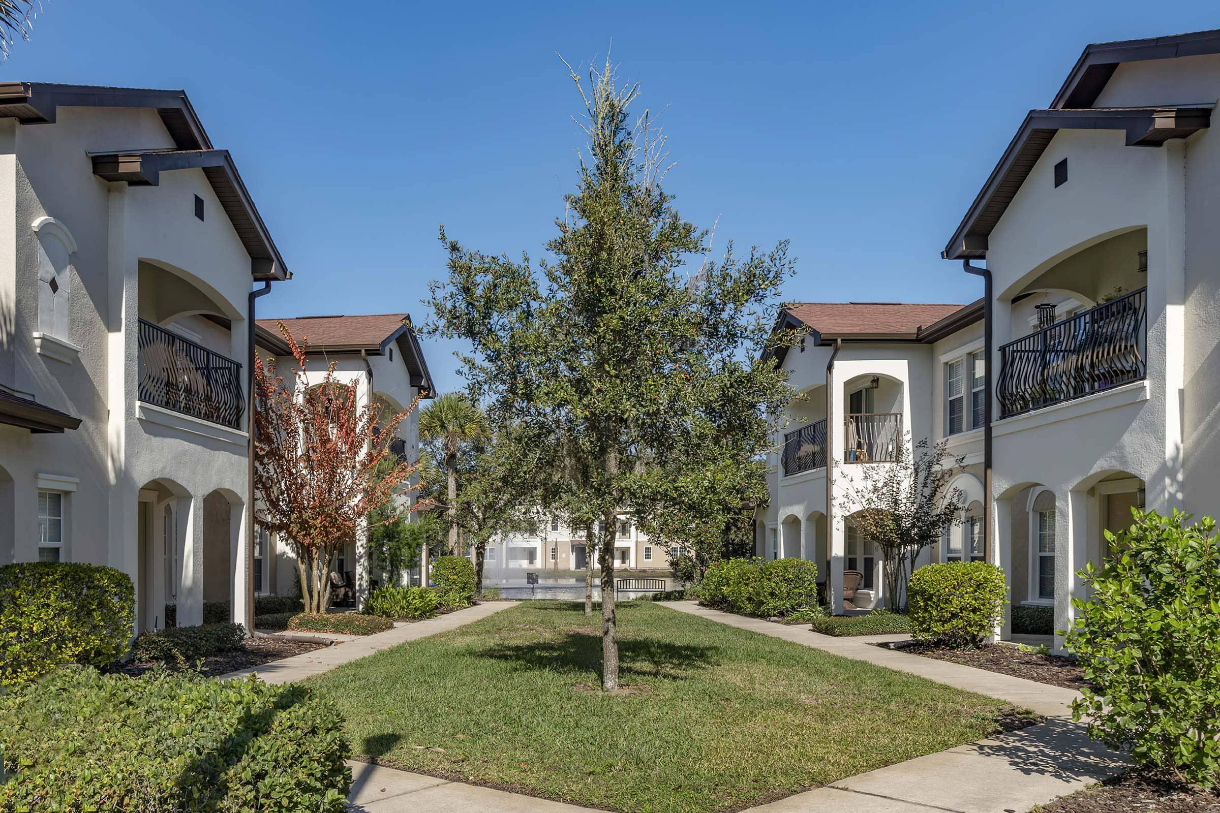 A well-maintained courtyard surrounded by two-story apartment buildings. The courtyard features a central tree, lush green grass, and neatly trimmed hedges. Bright, sunny sky with minimal clouds provides a cheerful atmosphere.