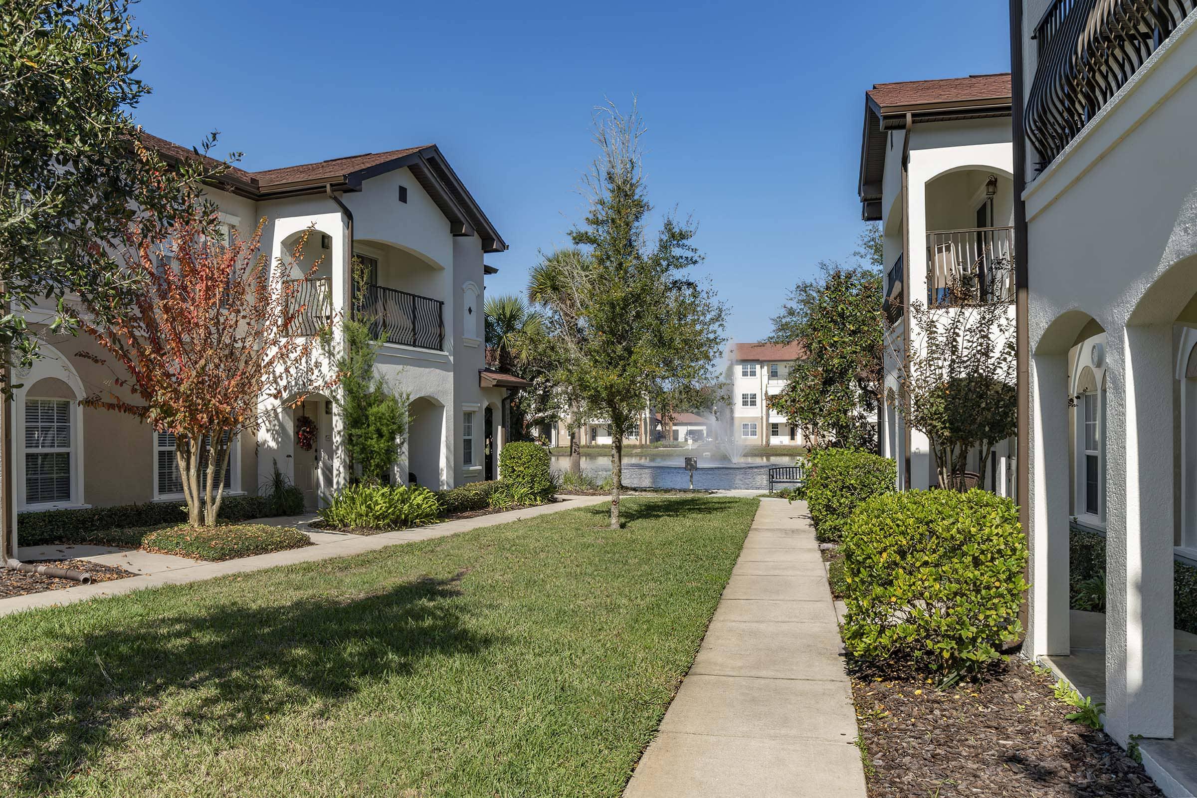 A tranquil courtyard scene featuring two multi-story apartment buildings on either side. The landscaped area includes neatly trimmed grass, bushes, and small trees. A pathway runs between the buildings, leading to a serene pond visible in the background under a clear blue sky.