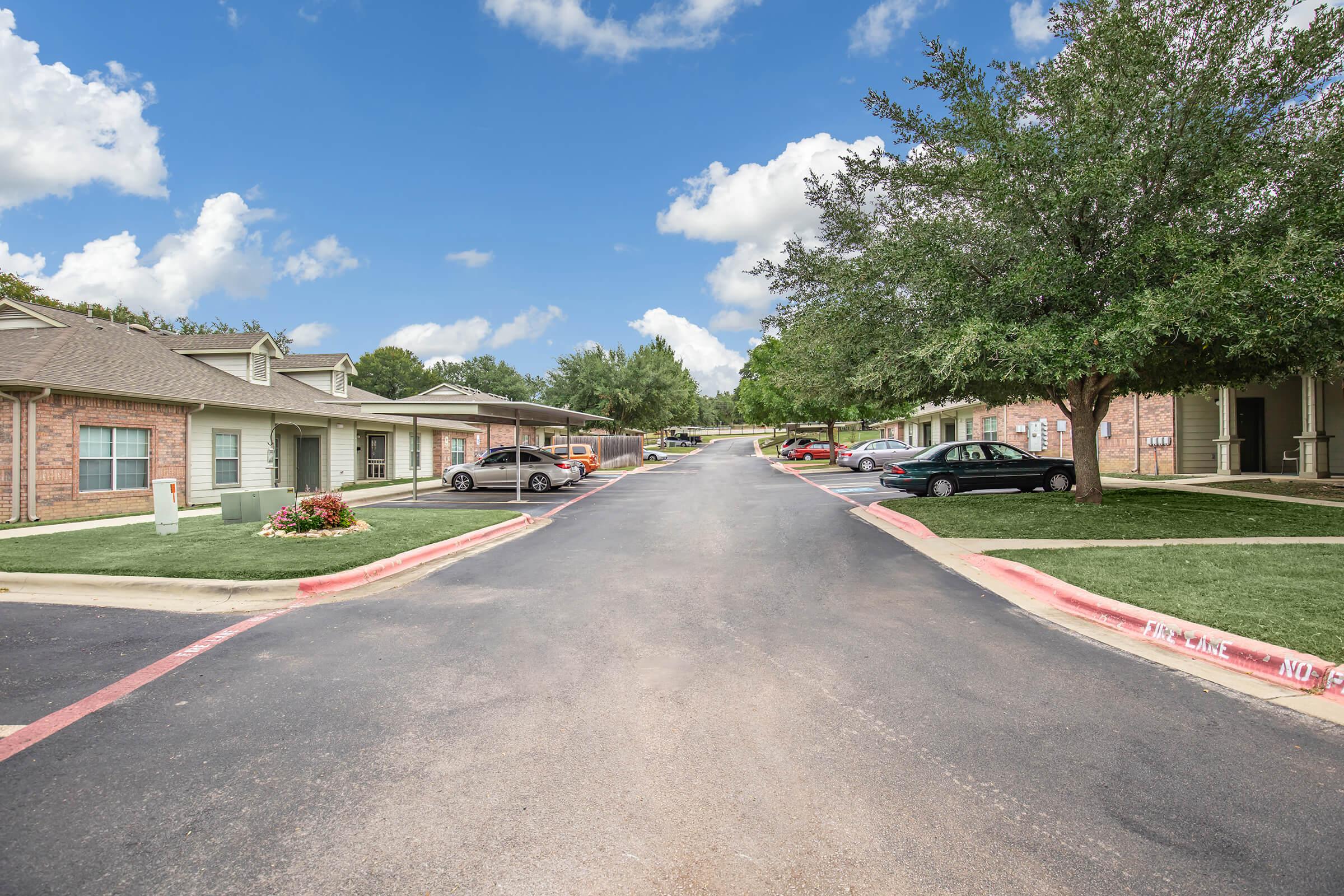 A landscaped residential area featuring low-rise brick buildings on both sides of a paved road. The scene includes neatly trimmed grass, several parked cars, and trees lining the street under a blue sky with fluffy white clouds.
