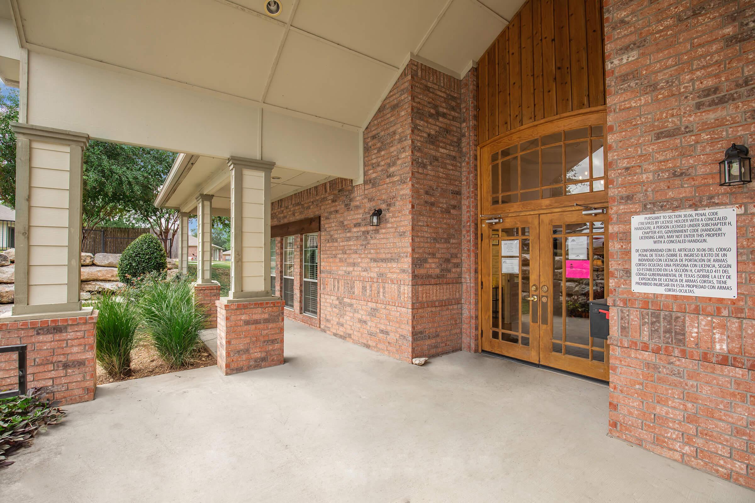 Exterior view of a brick building entrance with wooden double doors. The area features landscaped planters and a stone wall behind the entrance. There's a notice posted on the wall, and the surrounding space includes greenery and benches, creating an inviting atmosphere.