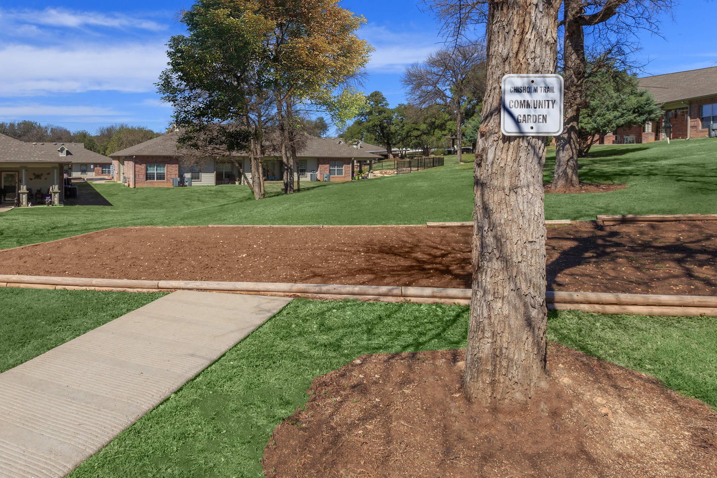 A community garden area marked by a sign on a tree, surrounded by green grass and trees. In the background, there are residential buildings with a clear blue sky above. The garden plot is empty, indicating it's ready for planting.