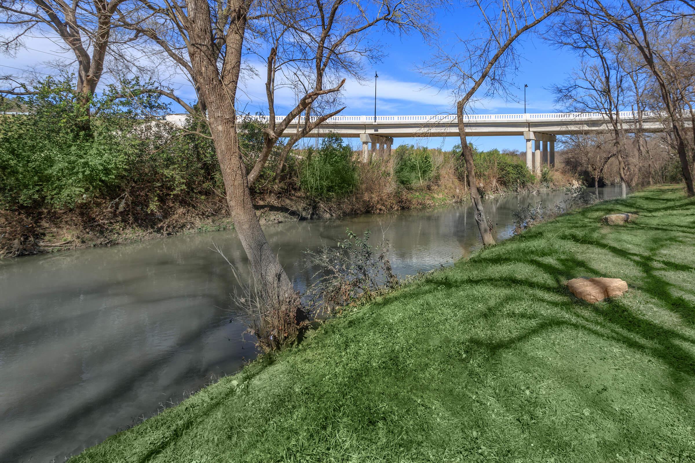 A serene river scene with a clear blue sky and scattered clouds. Lush green grass lines the bank, while trees with bare branches stand beside the water. A concrete bridge crosses over the river in the background, creating a tranquil natural setting.