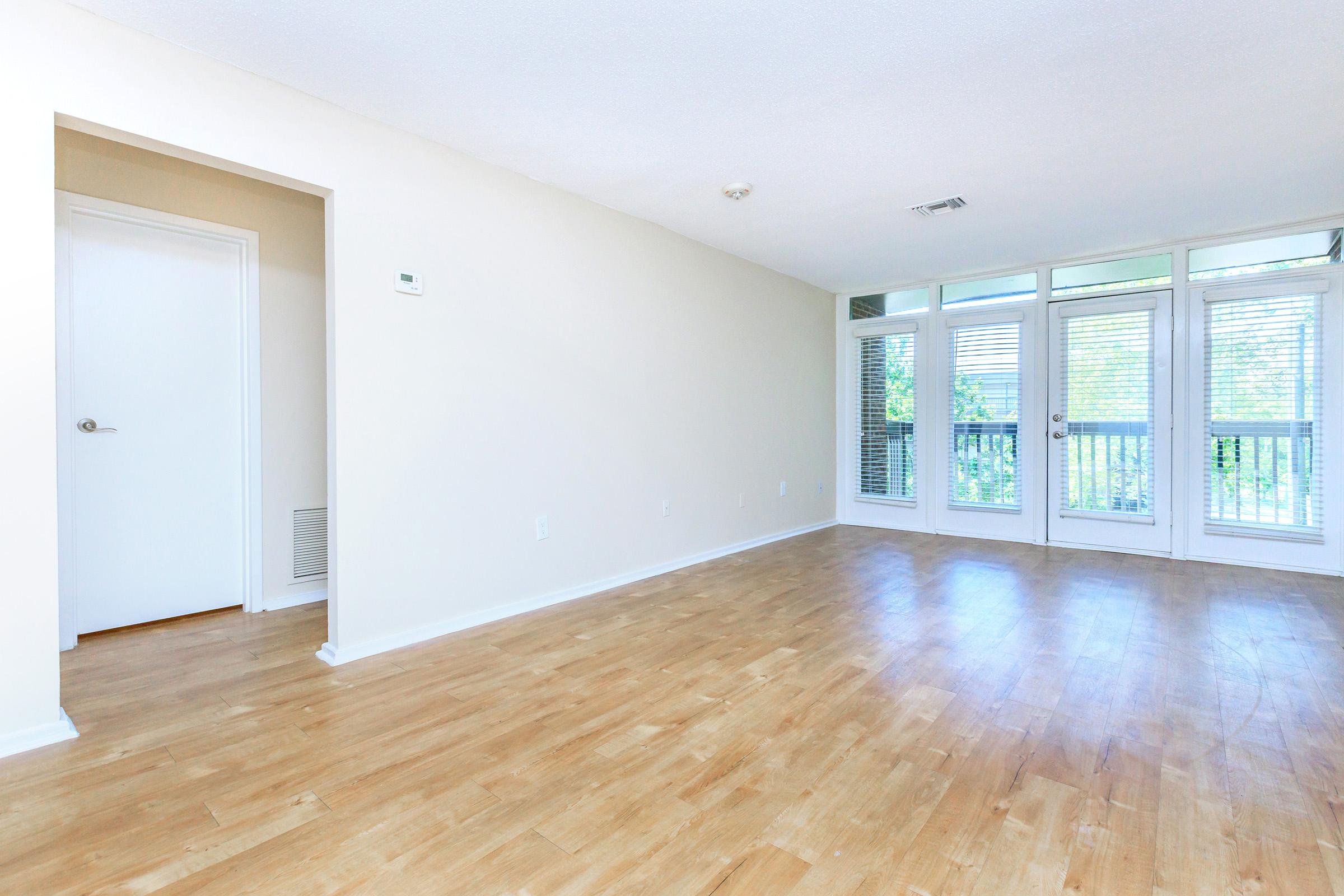 Empty room with light-colored walls and wooden flooring. There is a door on the left leading to another space and large windows on the right that provide natural light. The windows have blinds and overlook a balcony. The overall atmosphere is bright and spacious, creating an inviting environment.
