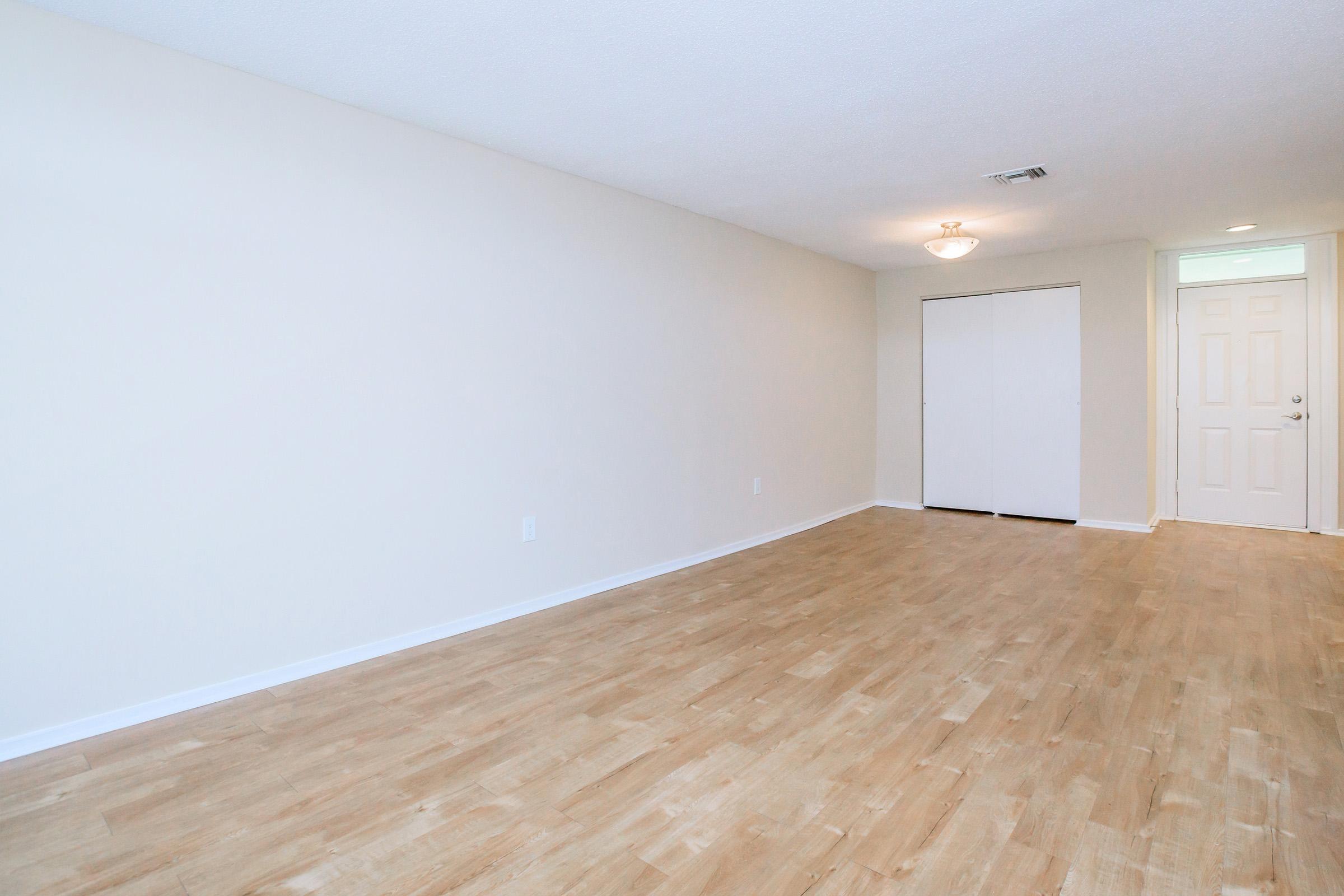 A spacious, empty room with light-colored walls and a wooden floor. There is a ceiling light fixture, and two closed white closet doors are visible on one side. A doorway leads to another area, suggesting a bright and open layout with natural light.