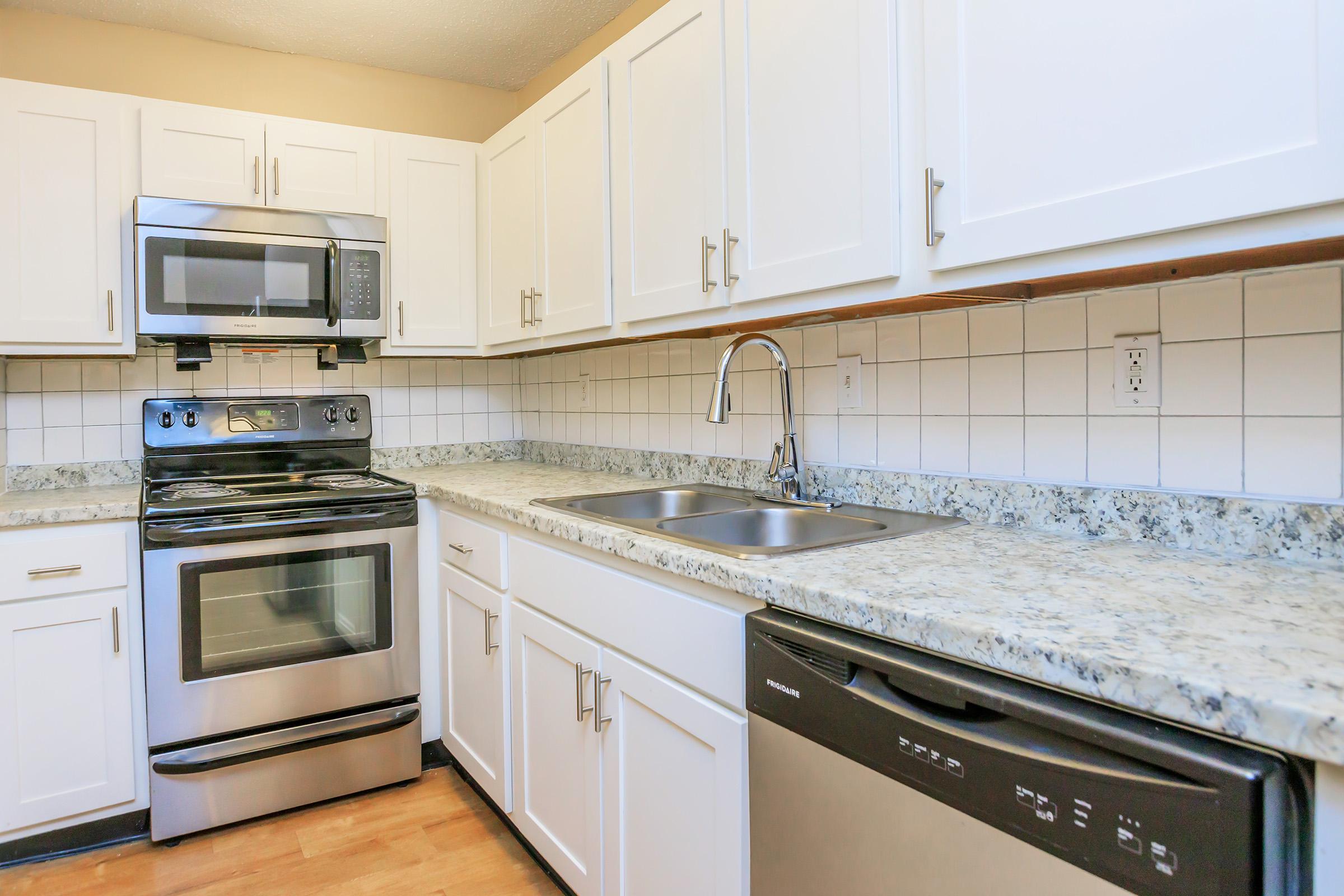 Modern kitchen featuring stainless steel appliances, including a microwave, oven, and dishwasher. White cabinets and a granite countertop complement the tiled backsplash. A double sink is installed under a sleek faucet, with wooden flooring enhancing the overall aesthetic.