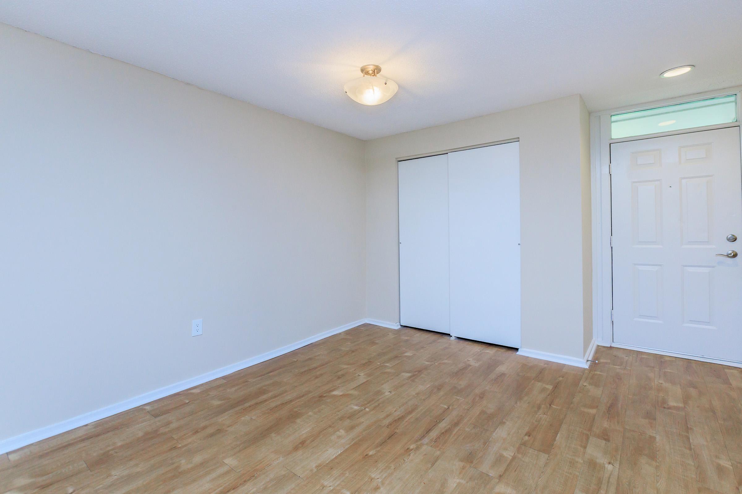 Empty room with light-colored walls and a wooden floor. One side features a double door closet, and the entrance door is visible on the right. A ceiling light fixture illuminates the space, creating a bright and clean atmosphere. No furniture or decorations are present, emphasizing the room's spaciousness.
