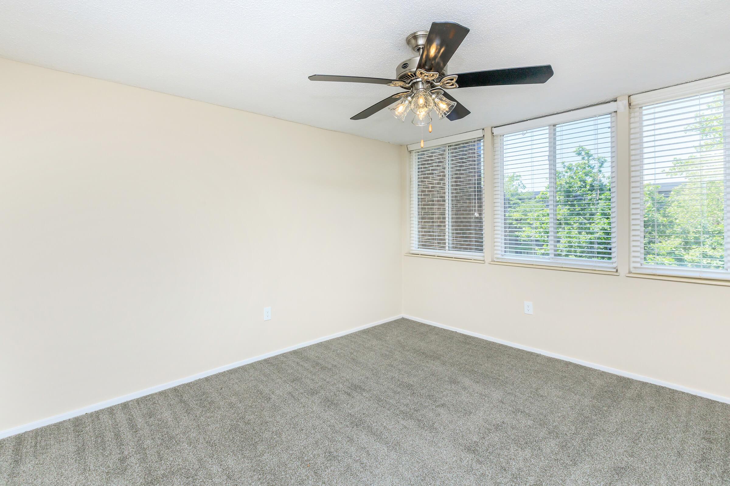 A light and airy room featuring beige walls and plush gray carpet. The space has a ceiling fan with a decorative light fixture and large windows with white blinds, allowing natural light to flood in and offering views of greenery outside.