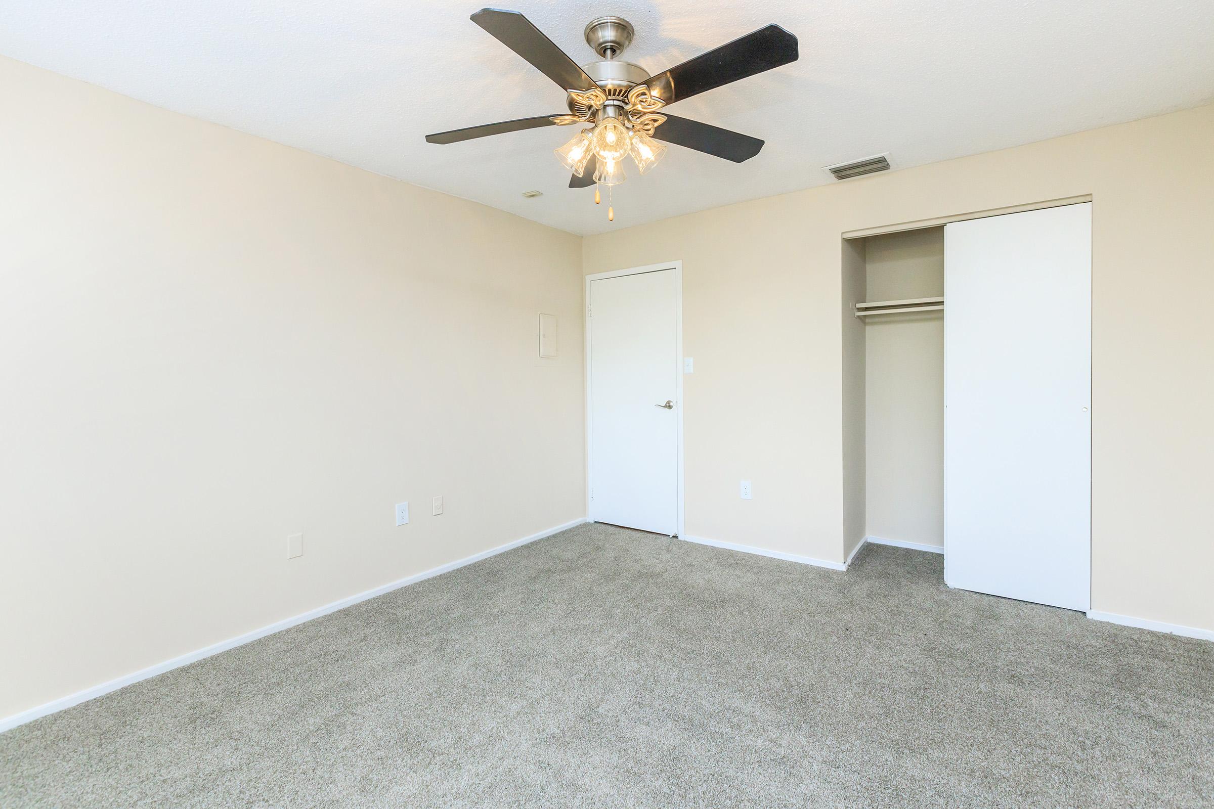 A spacious, empty room with beige walls and gray carpet. A ceiling fan with light is installed in the center. There is a closed white door on one wall and a sliding closet door on another, offering storage space. The room is well-lit and ready for furniture.