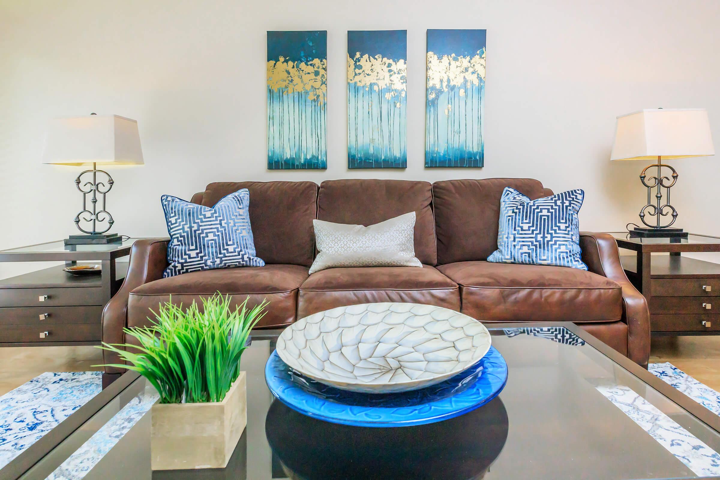 A cozy living room featuring a brown leather sofa adorned with decorative pillows. Two lamps flank the sofa on dark wooden side tables. Above the sofa are three abstract paintings in blue and gold. A stylish bowl sits on a blue plate in the center of a glass coffee table, complemented by a small greenery arrangement.
