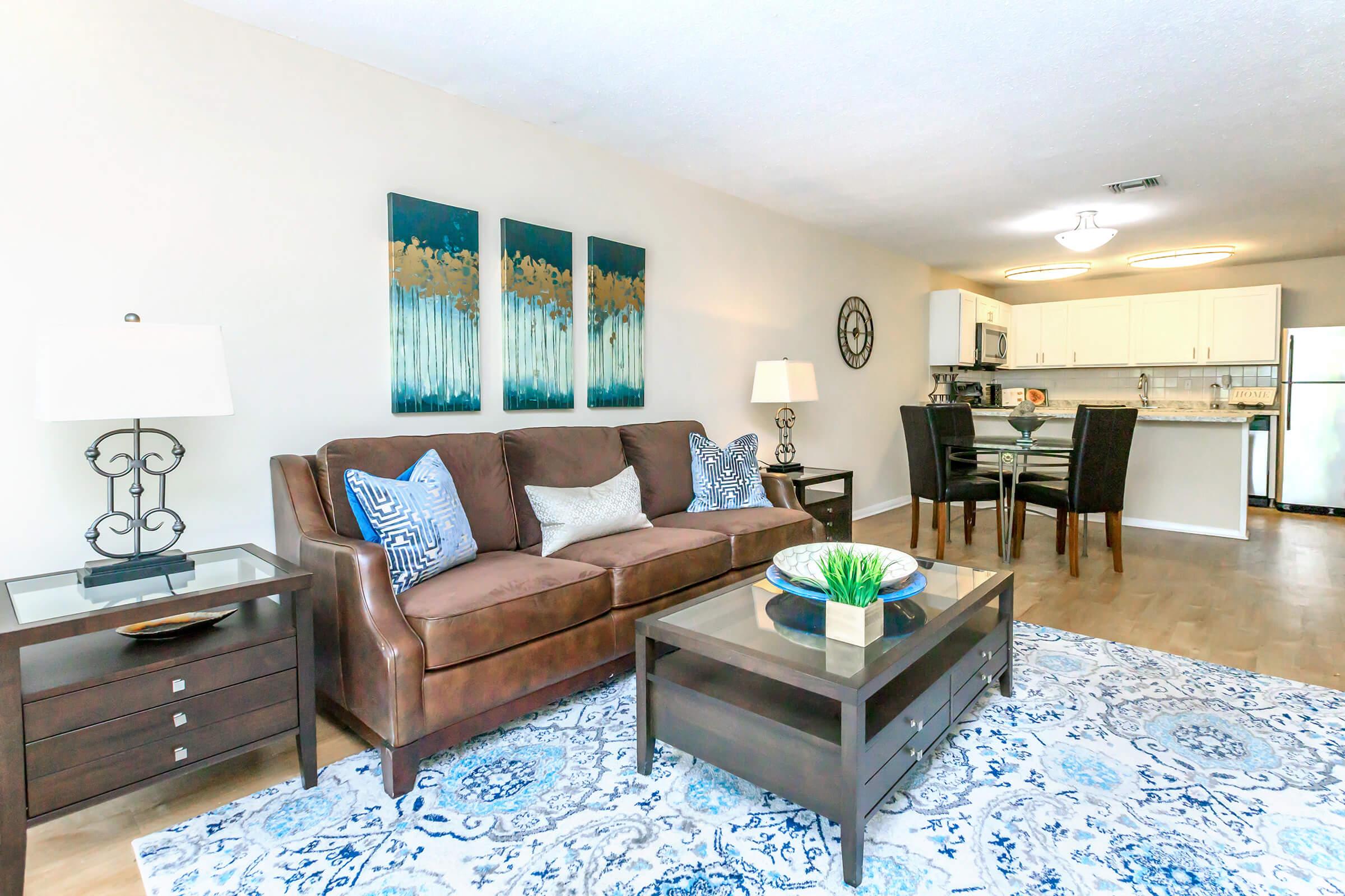 A cozy living area featuring a brown sofa with decorative pillows, two table lamps on side tables, and a blue and white area rug. The background shows a dining area with a glass table and chairs, along with a kitchen visible at the back, decorated with modern artwork on the wall.