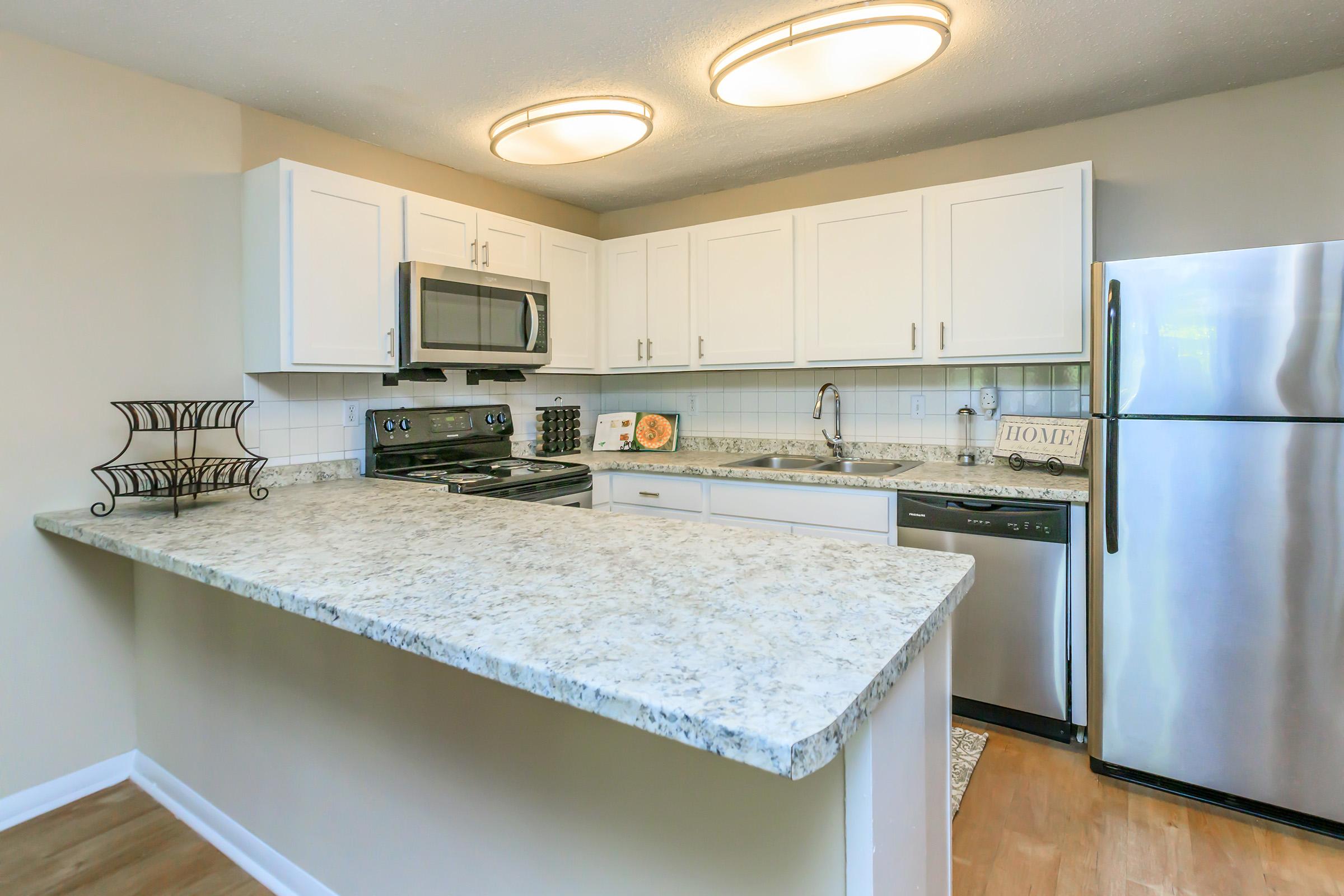 Modern kitchen featuring white cabinetry, stainless steel appliances, and granite countertops. A kitchen island with a granite surface separates the cooking area from the living space. Bright lighting fixtures illuminate the room, creating a warm and inviting atmosphere.