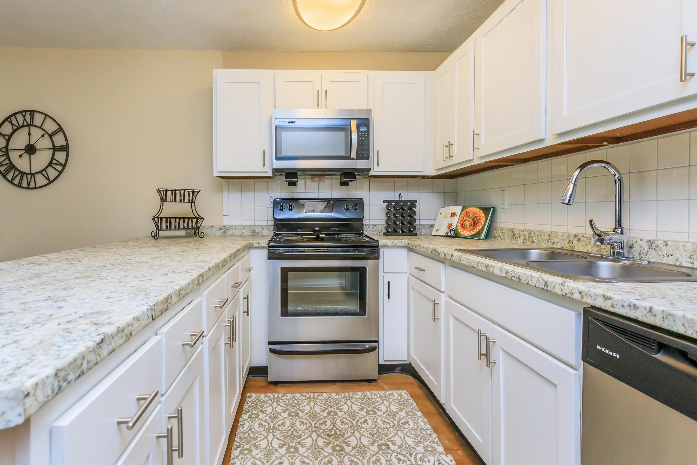A modern kitchen featuring white cabinets, stainless steel appliances, a granite countertop, and a double sink. A clock is mounted on the wall, and a decorative dish is displayed on the counter. The overall design is bright and clean, with a rug on the floor adding a cozy touch.