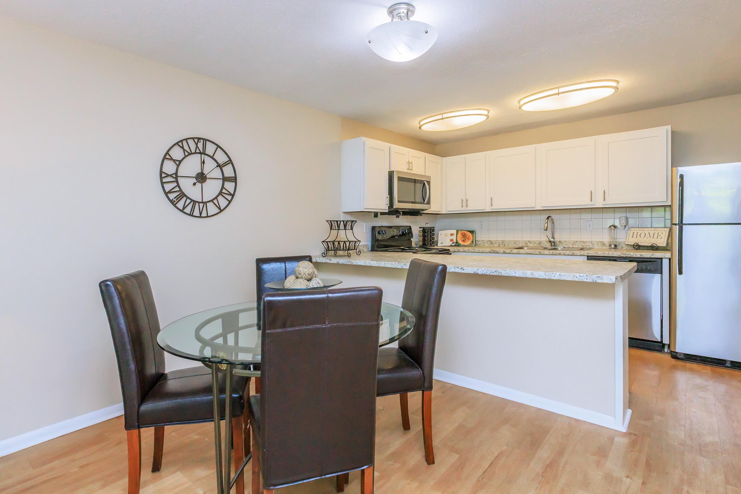 A modern kitchen and dining area featuring a glass dining table with four brown upholstered chairs, a wall clock, and a white kitchen with granite countertops. Appliances include a microwave, refrigerator, and stove, with light wood flooring creating a warm atmosphere.