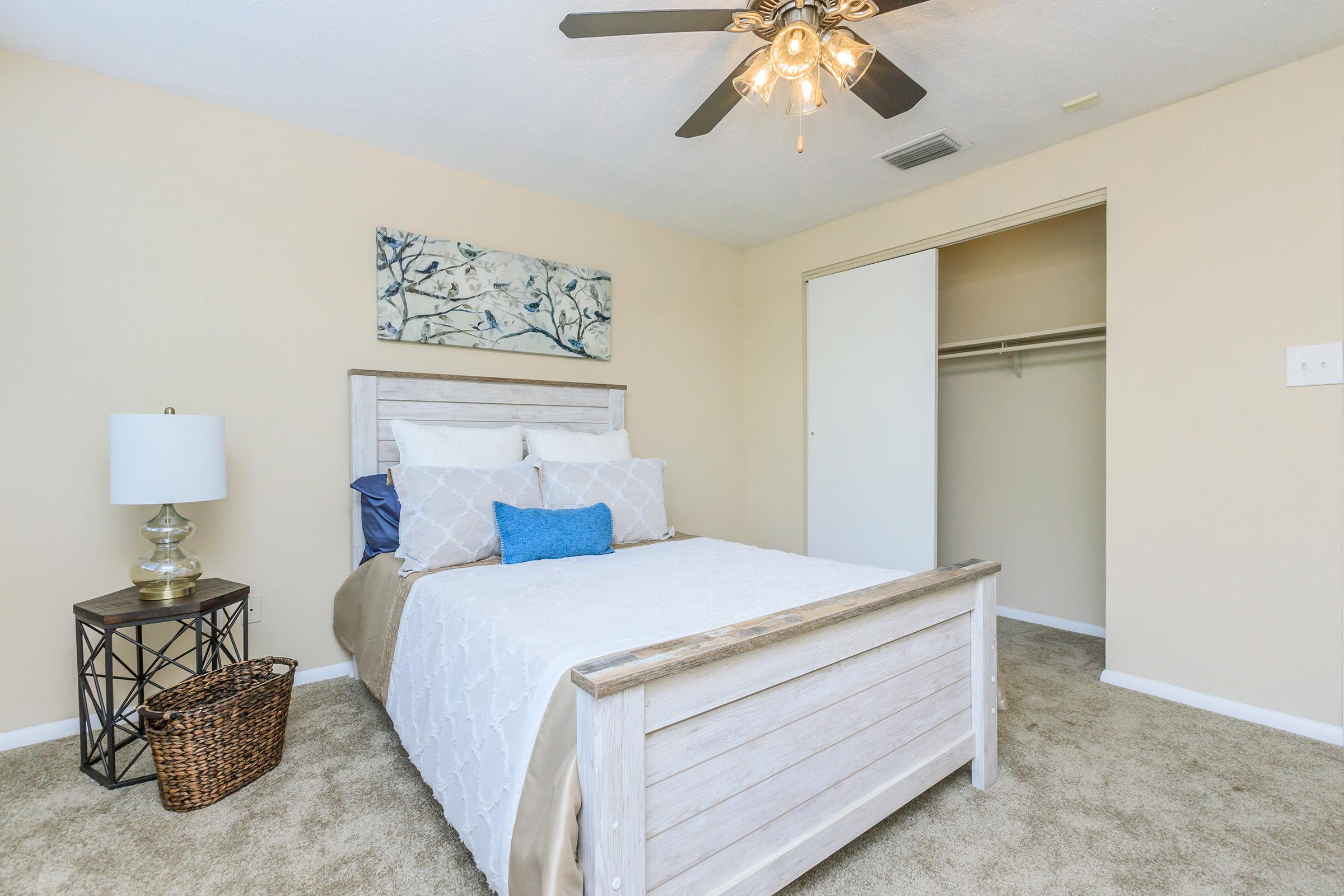 Cozy bedroom featuring a bed with white bedding and decorative blue pillows, a nightstand with a lamp, and a woven basket. The walls are a soft beige color, and there's an abstract artwork above the bed. A ceiling fan adds to the comfort, and a closet is visible in the background.