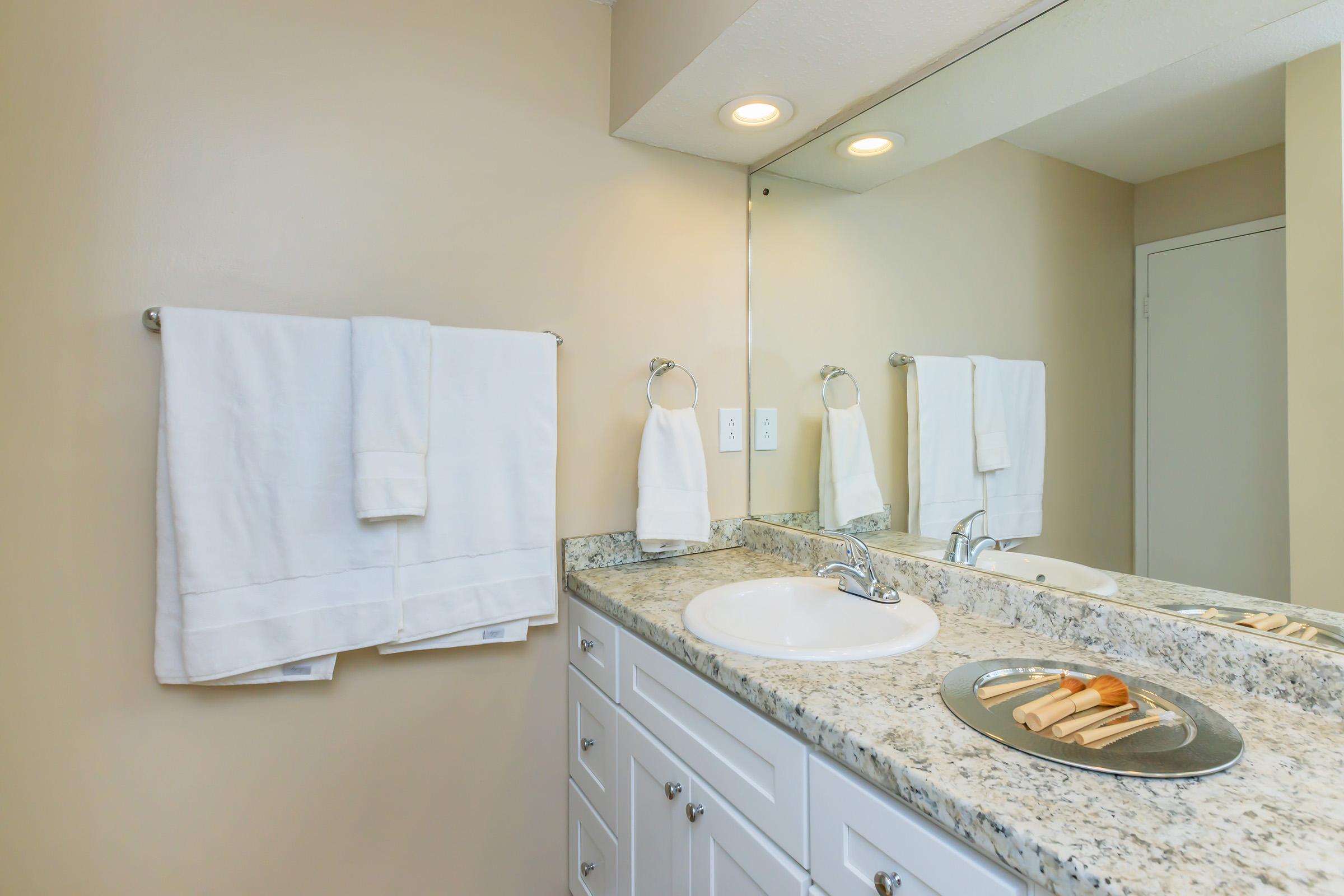 A clean bathroom with a light-colored wall and granite countertop. Two white towels hang on a towel rack. A sink with a faucet is visible, alongside a silver tray with wooden items. Soft overhead lighting illuminates the space, creating a fresh and welcoming atmosphere.