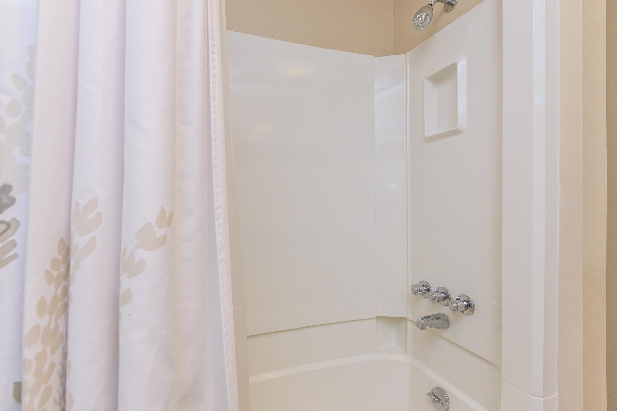 A clean shower area featuring a white tub with a showerhead and control knobs. A light-colored shower curtain hangs partially closed, and the walls are tiled with a smooth surface. The overall setting is bright and minimalist, conveying a sense of openness and hygiene.