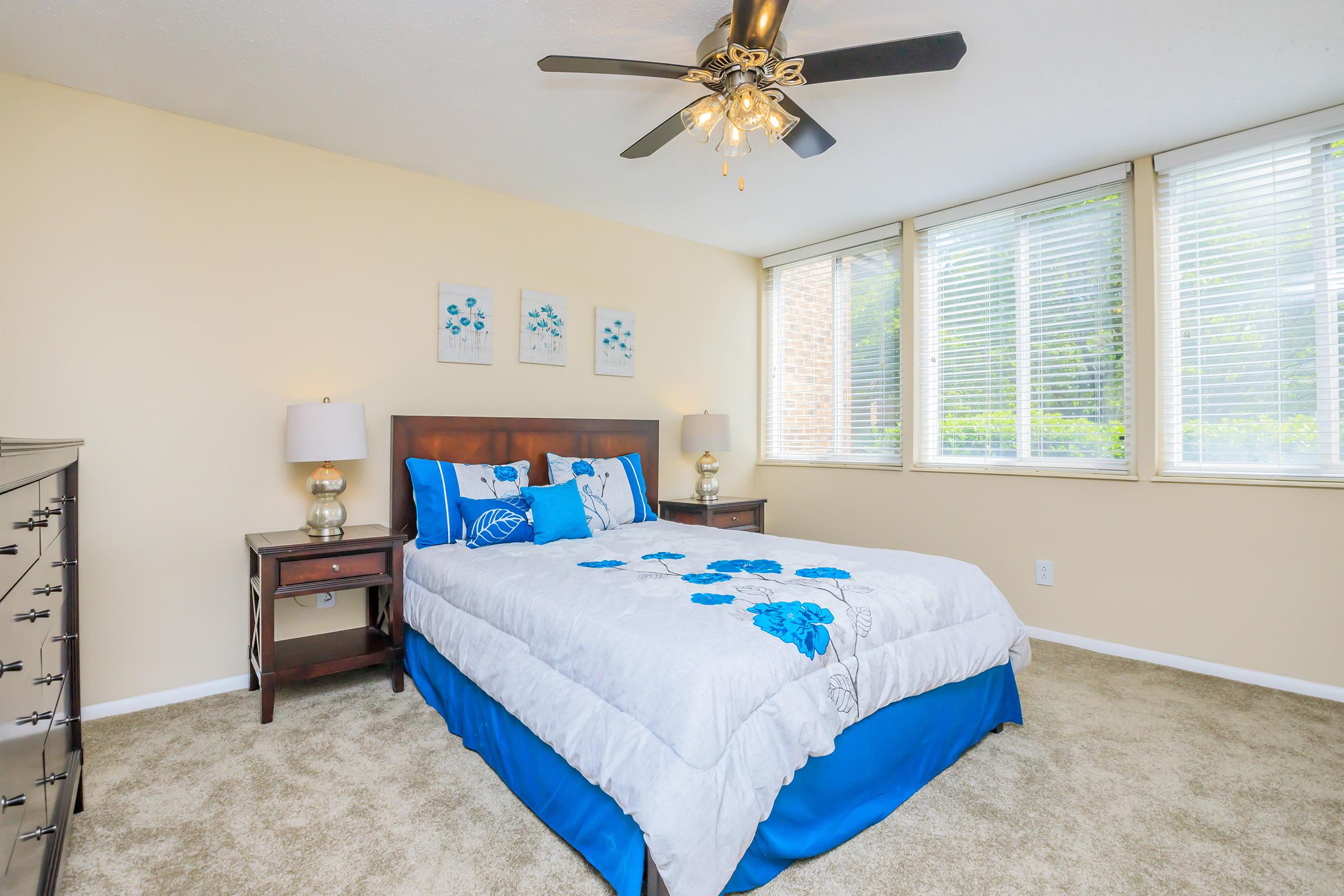 A bright and inviting bedroom featuring a queen-sized bed with blue and white floral bedding. Two nightstands with lamps flank the bed, and a dresser is positioned nearby. Large windows fill the room with natural light, and soft beige walls complement the cozy carpet.
