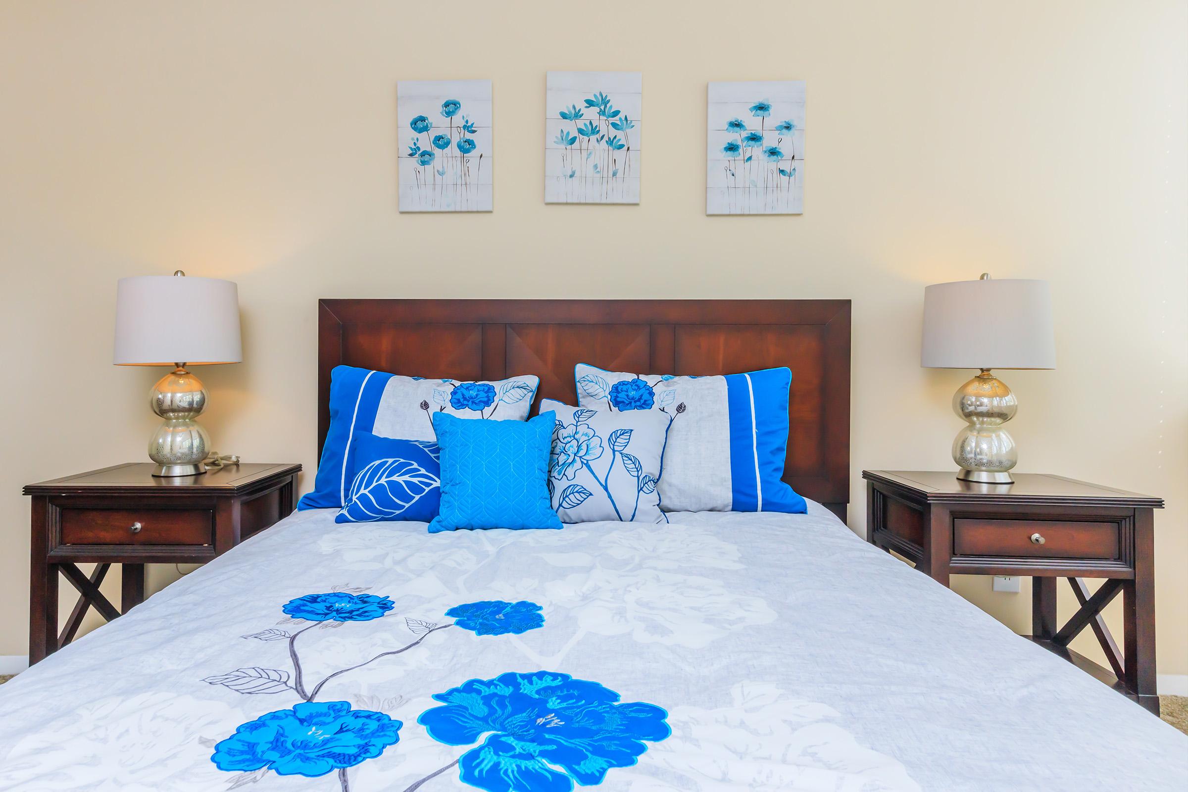 A cozy bedroom featuring a wooden bed frame with a white and blue floral comforter. Two decorative pillows add a pop of color. Flanking the bed are stylish nightstands with silver lamps. Above the bed hang three framed floral artworks, enhancing the serene atmosphere of the room. The walls are painted a soft beige.
