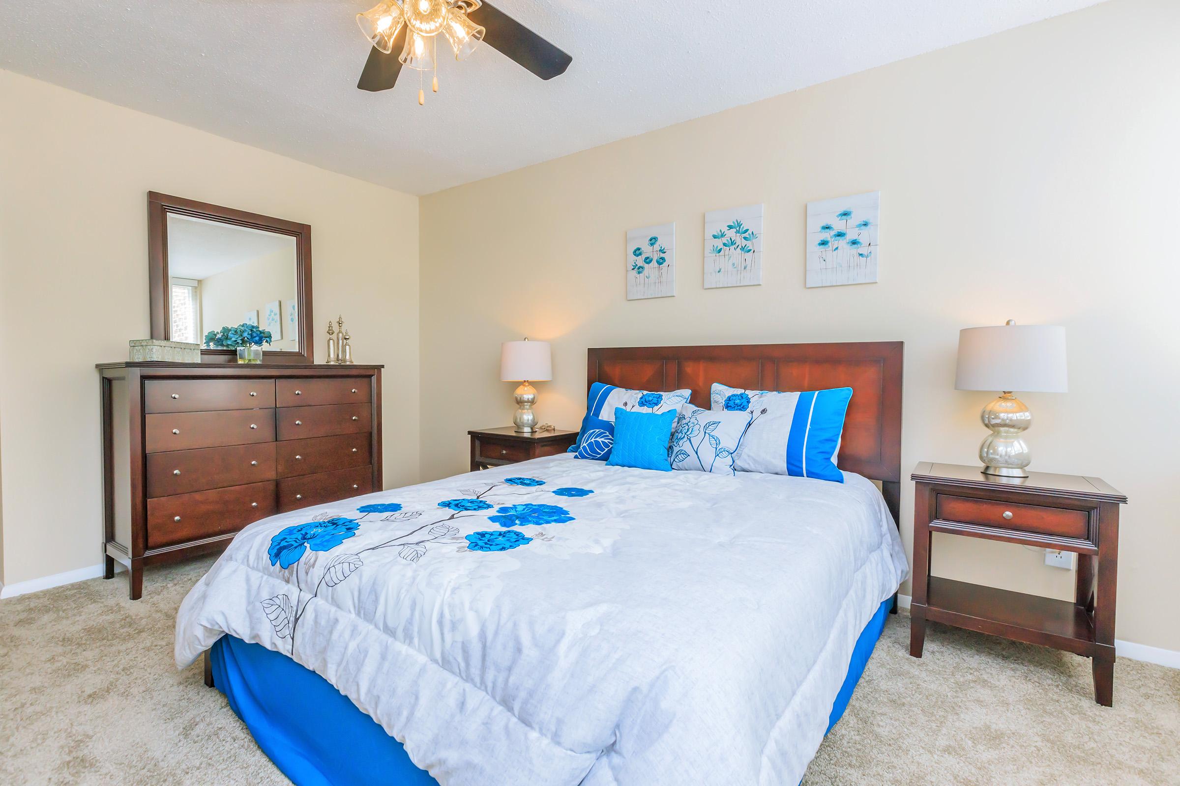 A cozy bedroom featuring a queen-sized bed with a blue and gray floral comforter, matching decorative pillows, and two bedside tables with lamps. A wooden dresser is visible, along with artwork on the wall, and a ceiling fan adds to the ambiance. The carpeted floor complements the light-colored walls.