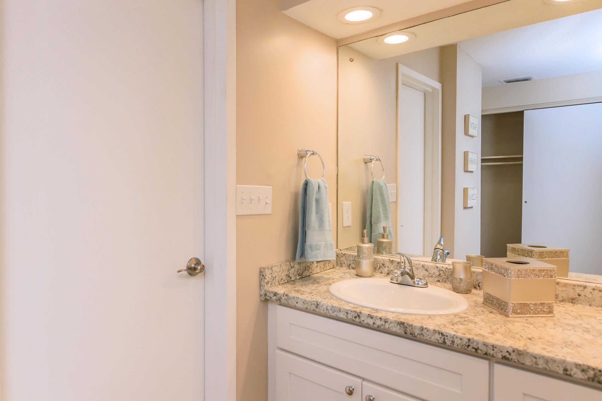 A clean bathroom featuring a white sink with a granite countertop, a large mirror above, light-colored walls, and a closed white door. Two hand towels hang on the wall, and decorative items are neatly arranged on the counter, creating a serene and organized atmosphere.