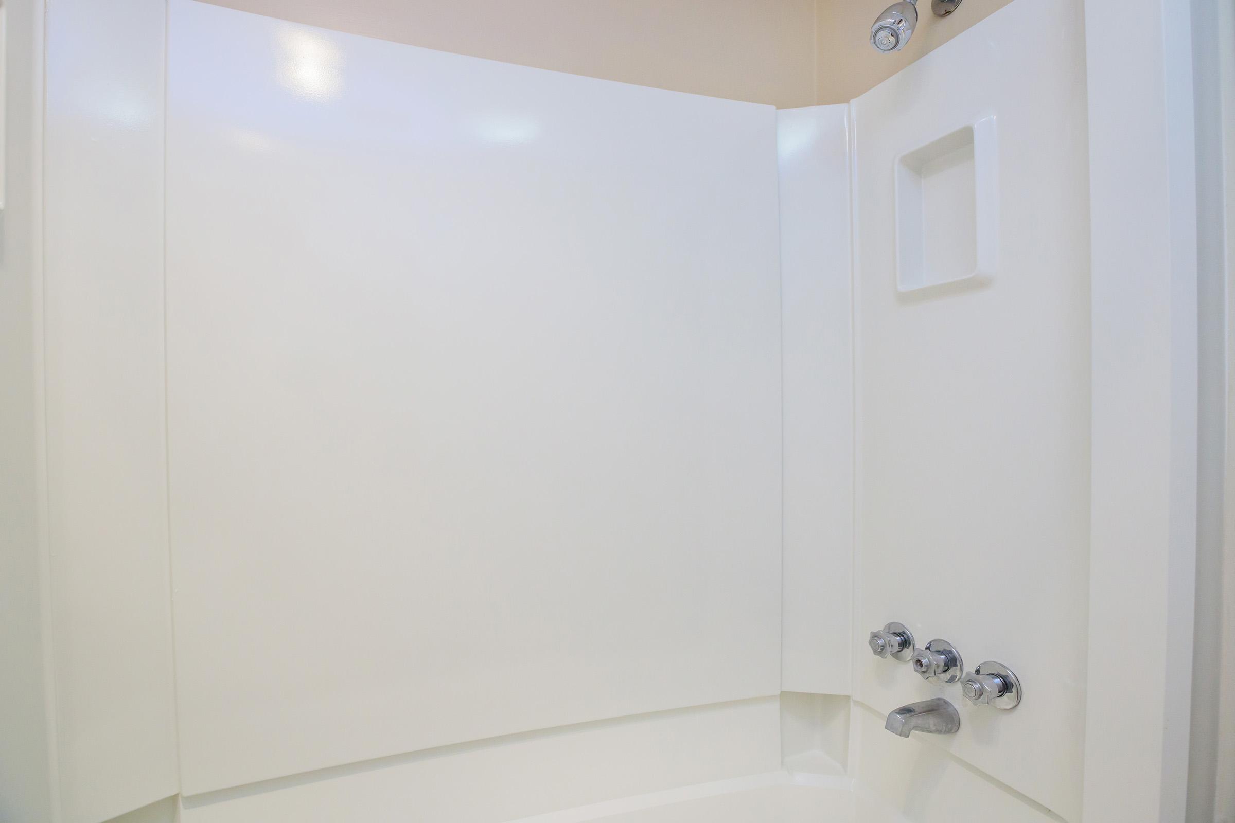A clean and modern shower stall with a white surround, featuring a built-in shelf and chrome fixtures. The design is minimalist, emphasizing a bright and welcoming bathroom space.