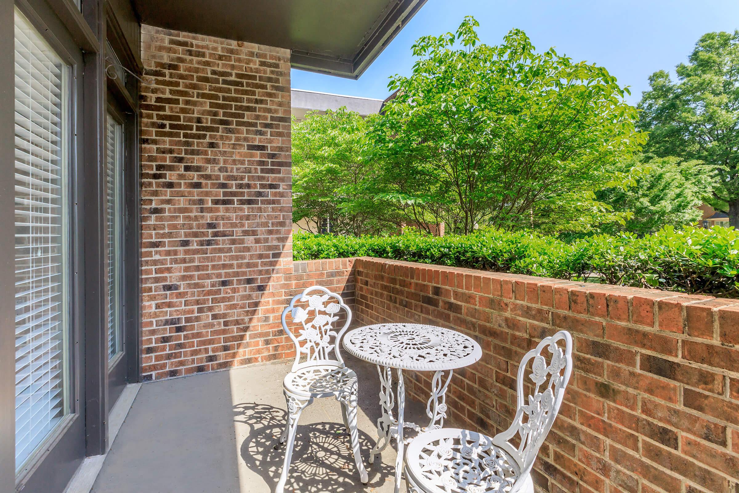 A small outdoor balcony featuring a round white table and two decorative chairs, surrounded by a brick wall and lush greenery in the background. Sunlight brightens the space, creating a cozy and inviting atmosphere.
