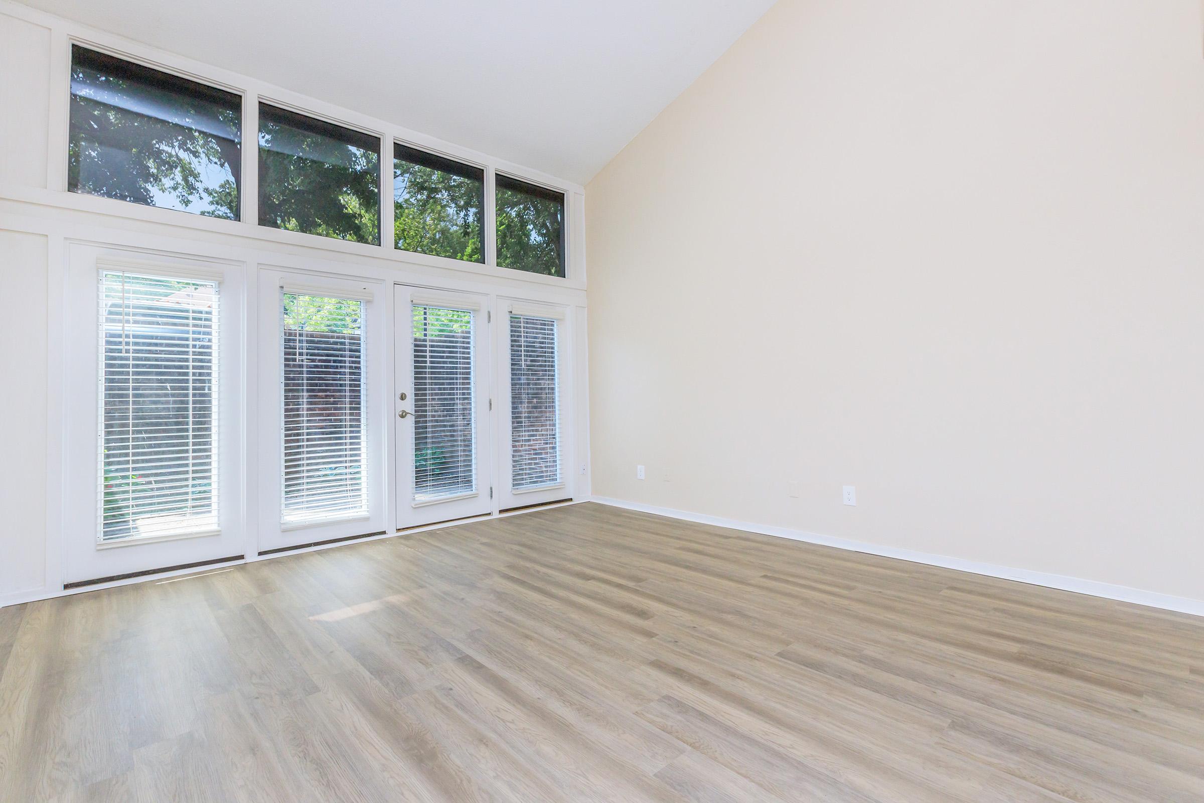 Spacious, empty room with high ceilings and large windows allowing natural light. The floor is made of light-colored wood laminate, and there are sliding glass doors leading to an outdoor area. The walls are painted a soft, neutral color, providing a blank canvas for decoration.