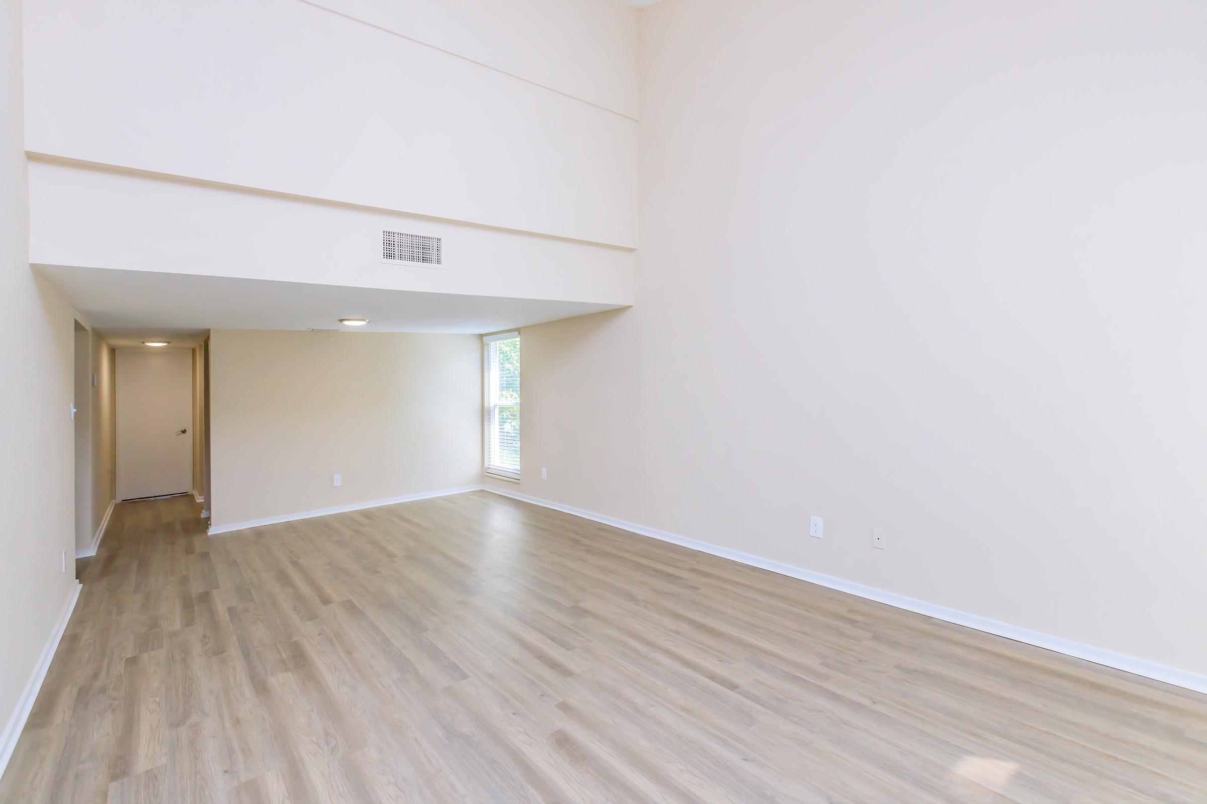Spacious, empty room with light beige walls and wooden flooring. Large windows allow natural light to fill the space. The ceiling is high, enhancing the feeling of openness. A doorway visible in the background leads to another area. The overall atmosphere is bright and airy.