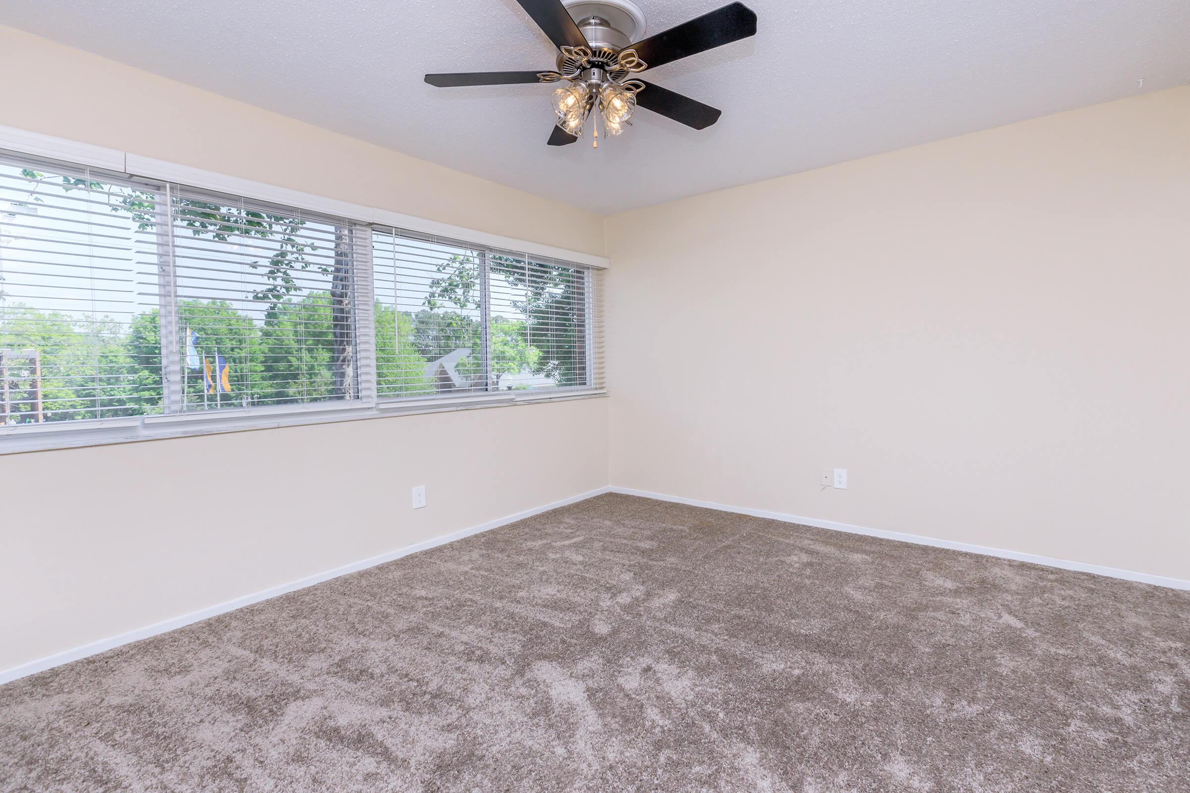A spacious, empty room with light beige walls, a ceiling fan with lights, and a large window showcasing greenery outside. The floor is covered in soft, light brown carpet, creating a warm and inviting atmosphere.