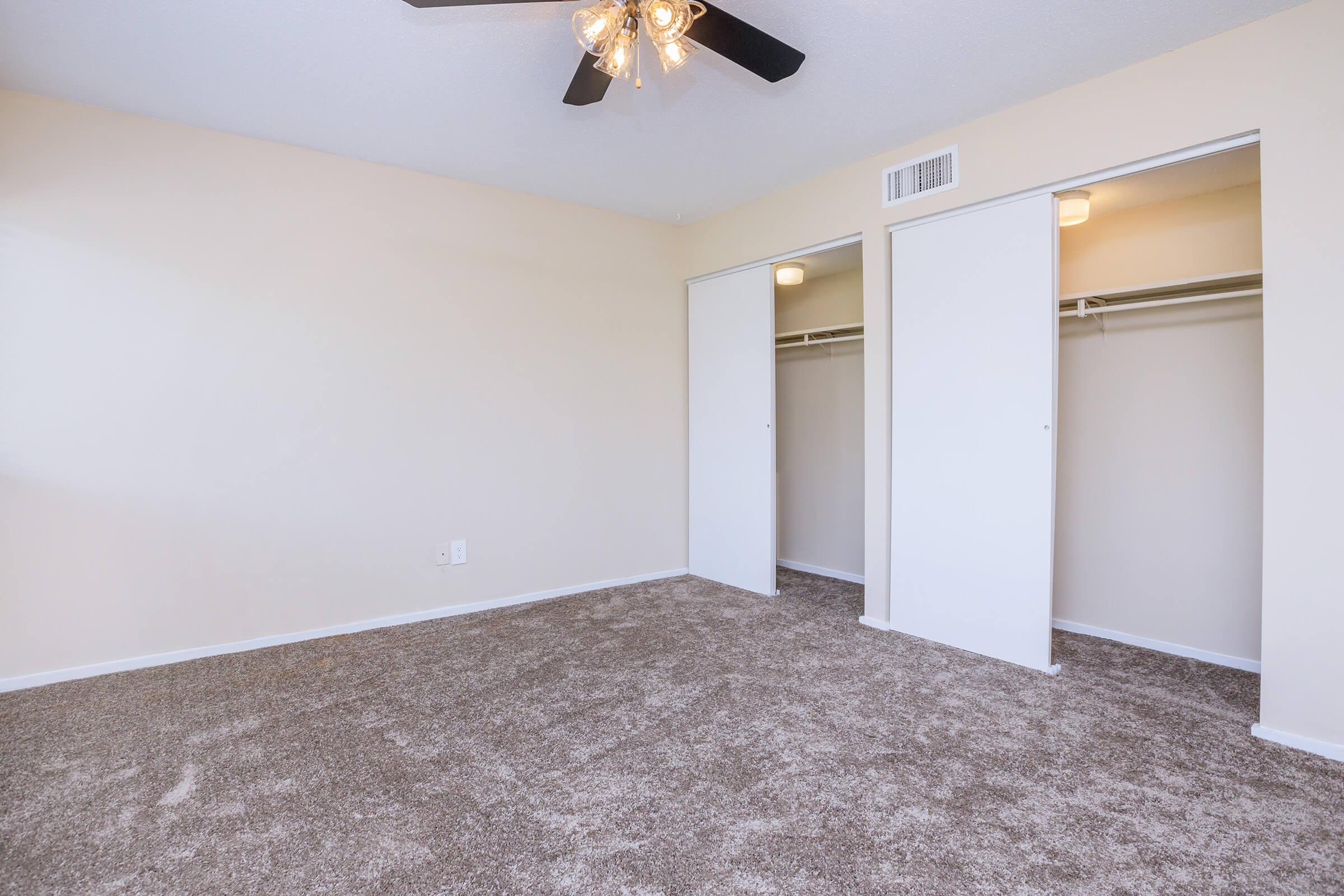 A spacious, empty room with light beige walls and a ceiling fan. The floor is covered in plush, light brown carpet. There are two closet doors on the right side, leading to ample storage space, and a single light fixture in the ceiling. The overall atmosphere is bright and inviting.